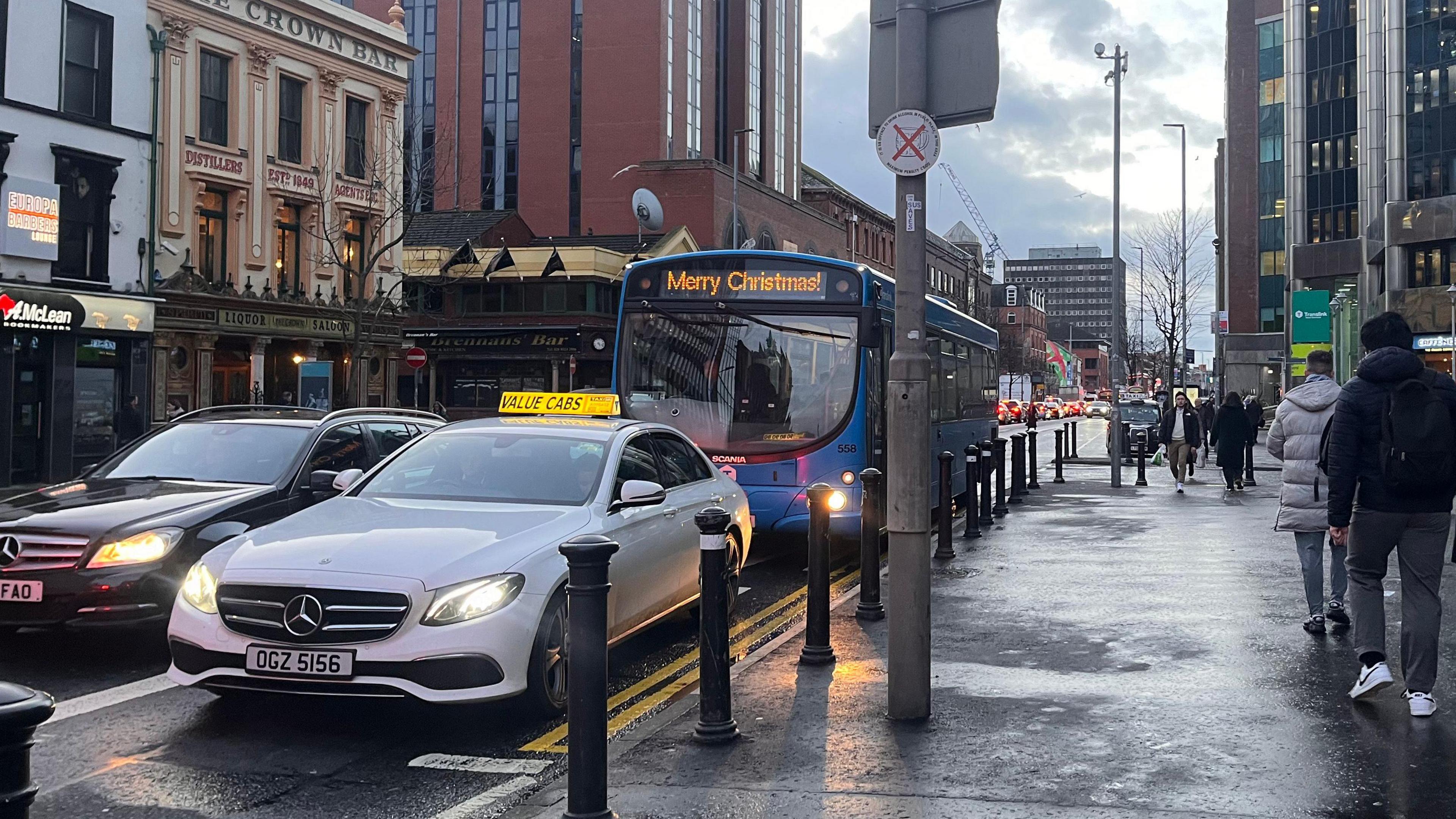 A while Mercedes Value Cabs taxi driving in a bus lane on Belfast's Great Victoria Street before Christmas.  A bus with a sign that says "Merry Christmas" is behind the taxi.