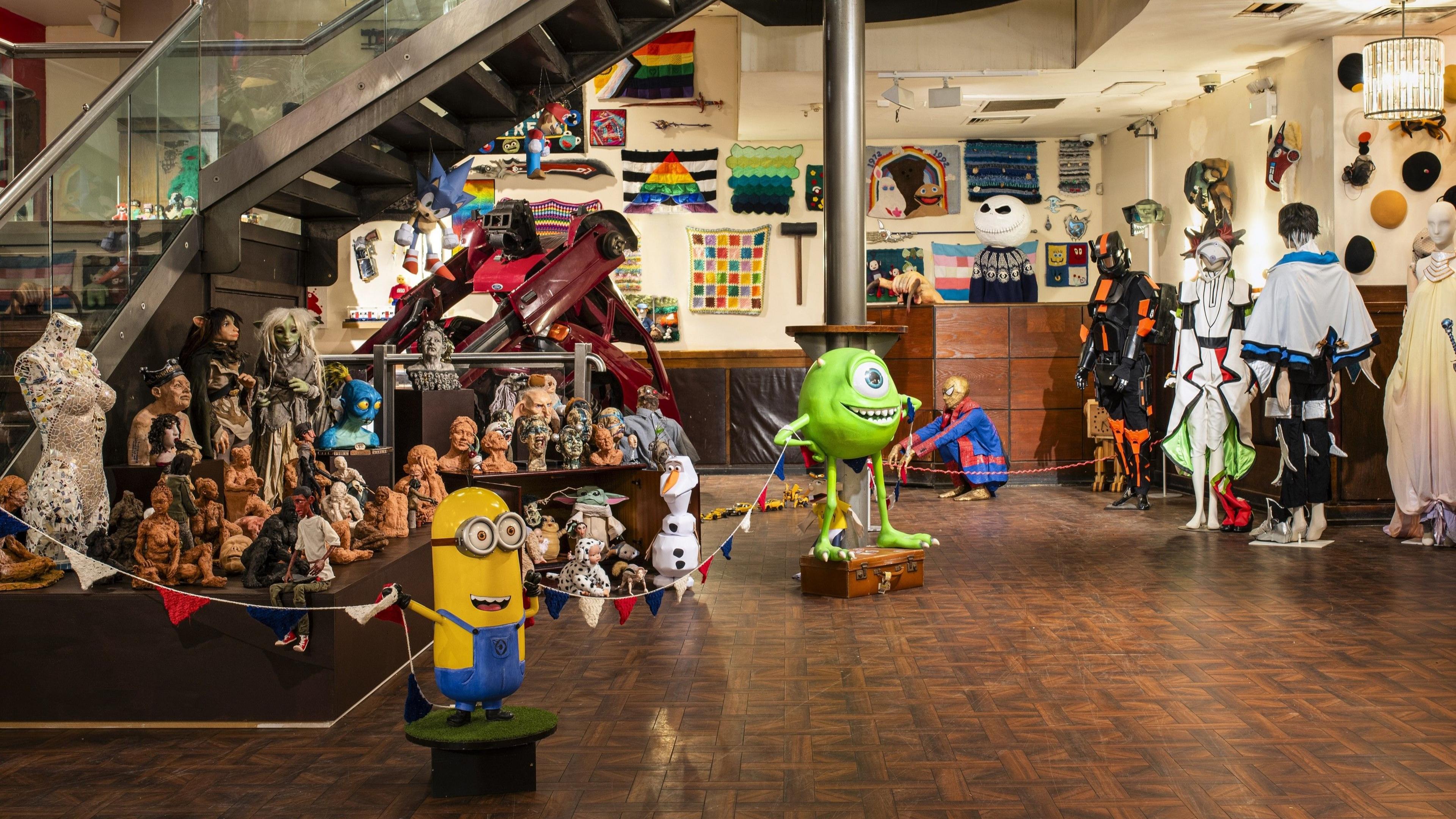 A room full of various items donated by hobbyists. There is a large yellow Minion figurine and a large Mike Wazowski figure from Monsters Inc, which is bright green. Behind them are costumes on mannequins and various small sculptures of busts or people.