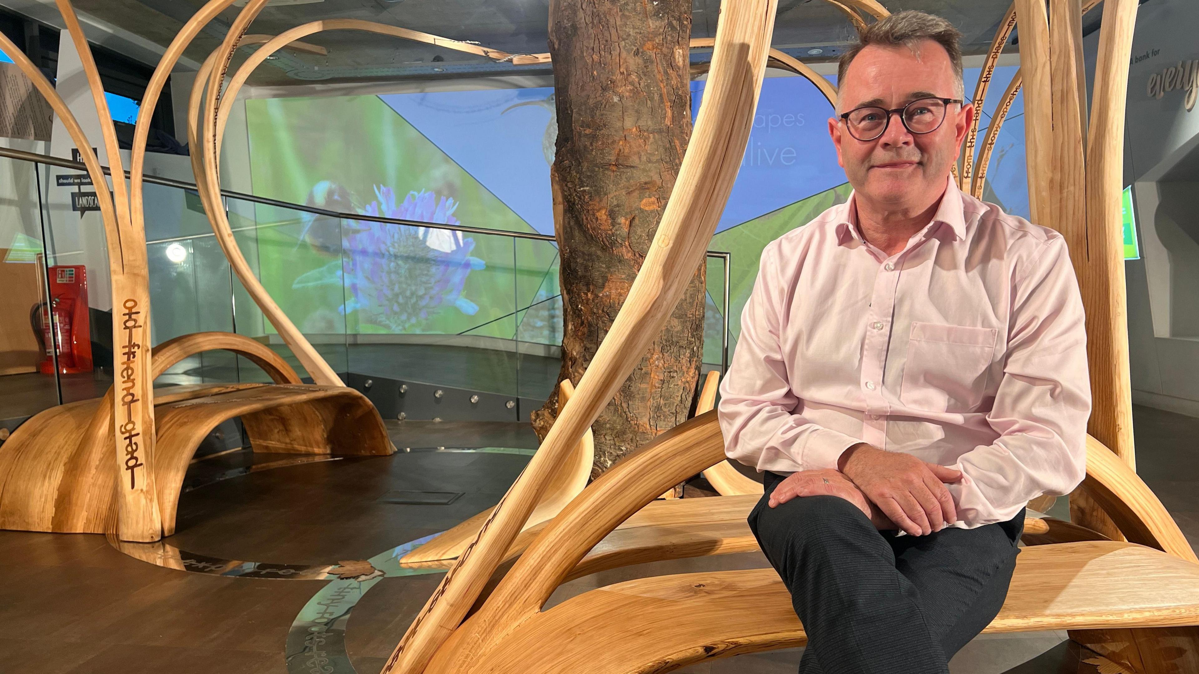 Tony Gates sits on a bench next to the trunk. He is clean shaven with short greying hair and is wearing glasses and a pink shirt.
