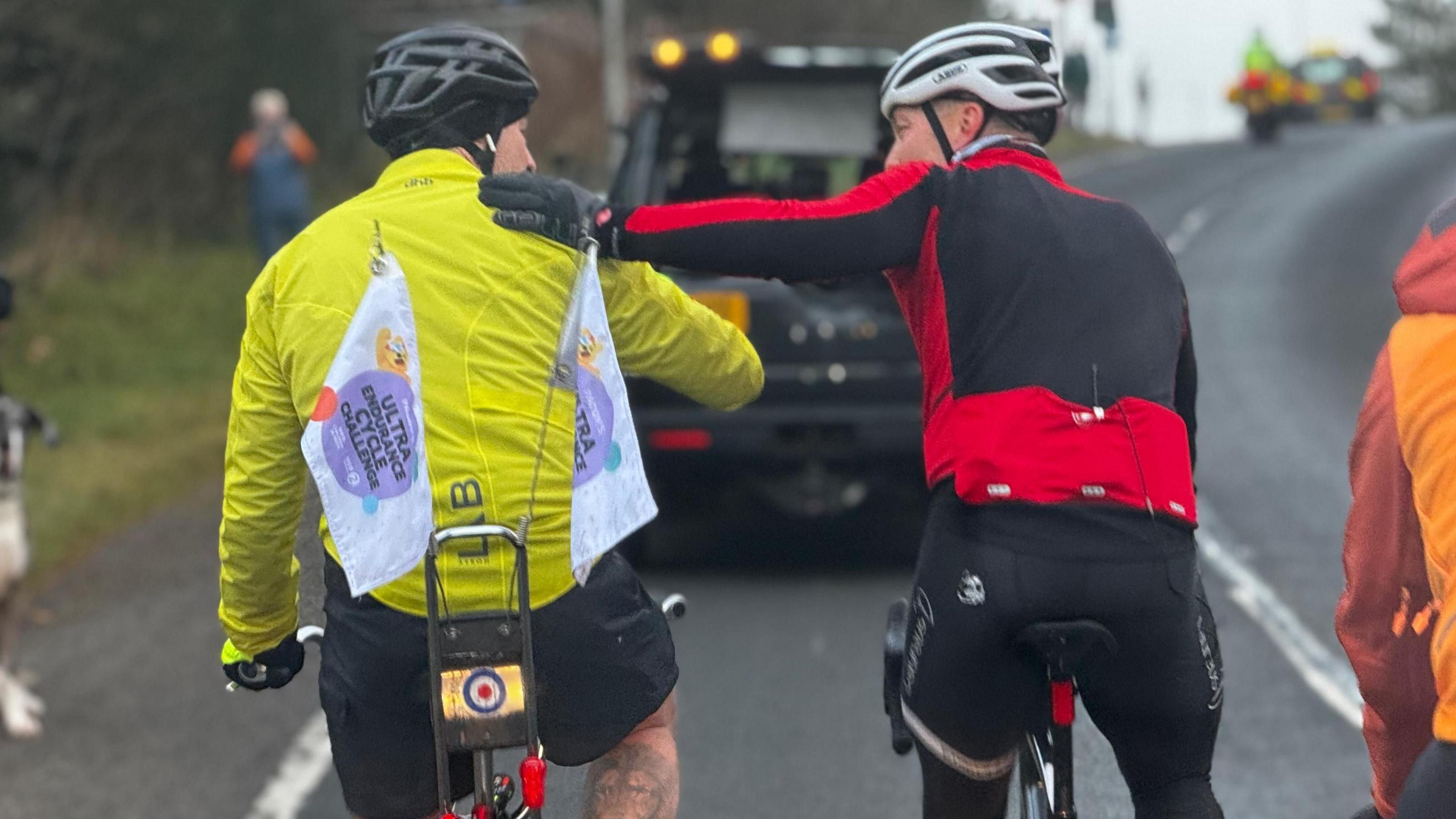 Photo from behind with Paddy McGuinness on the left, riding his chopper and wearing a bright hi-vis jacket with Children in Need banners flying from the back of the bike with Sir Chris Hoy on the right wearing a black and red jacket, his left arm outstretched and touching Paddy's shoulder as they cycle together on a road