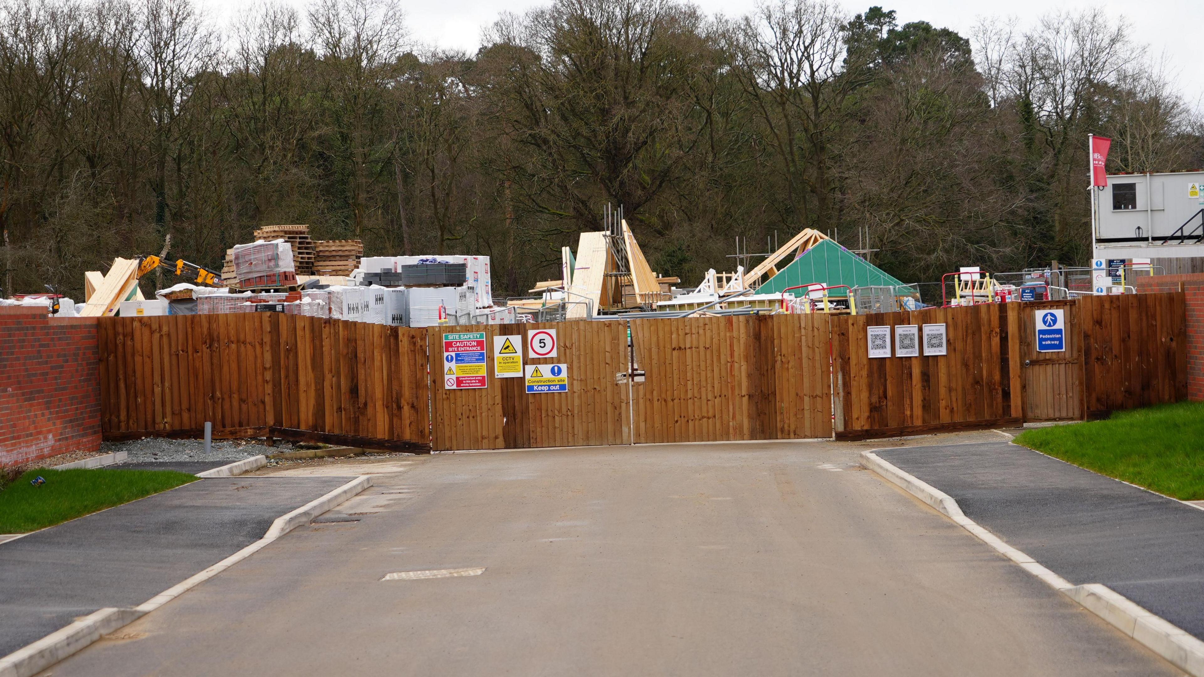 Tall wooden slat fencing, with construction notices, runs across a road. There are pallets and building materials behind the fencing.