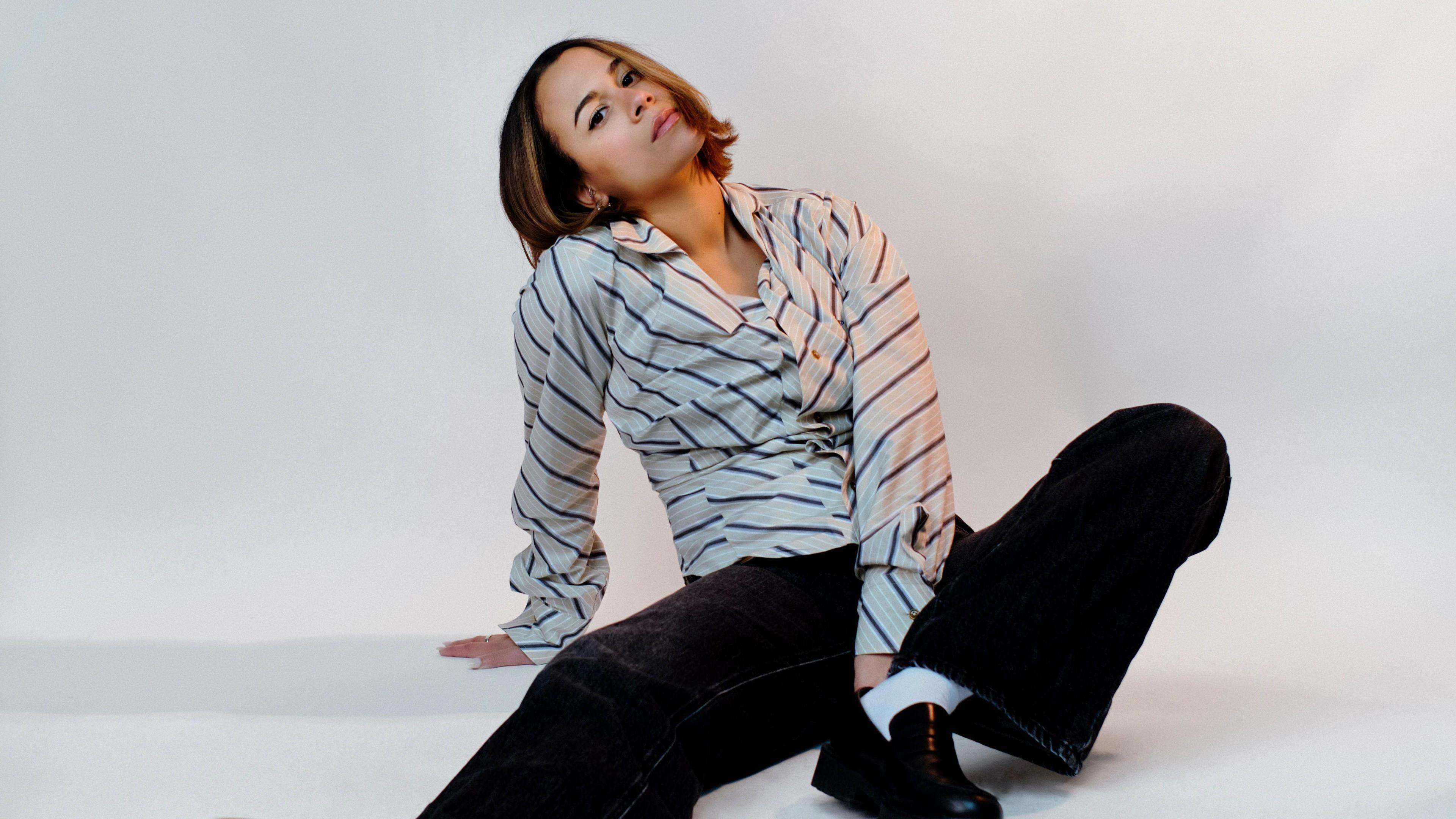 Alice Dallin in black trousers and a white , striped top, sitting on the floor while holding one of her knees up