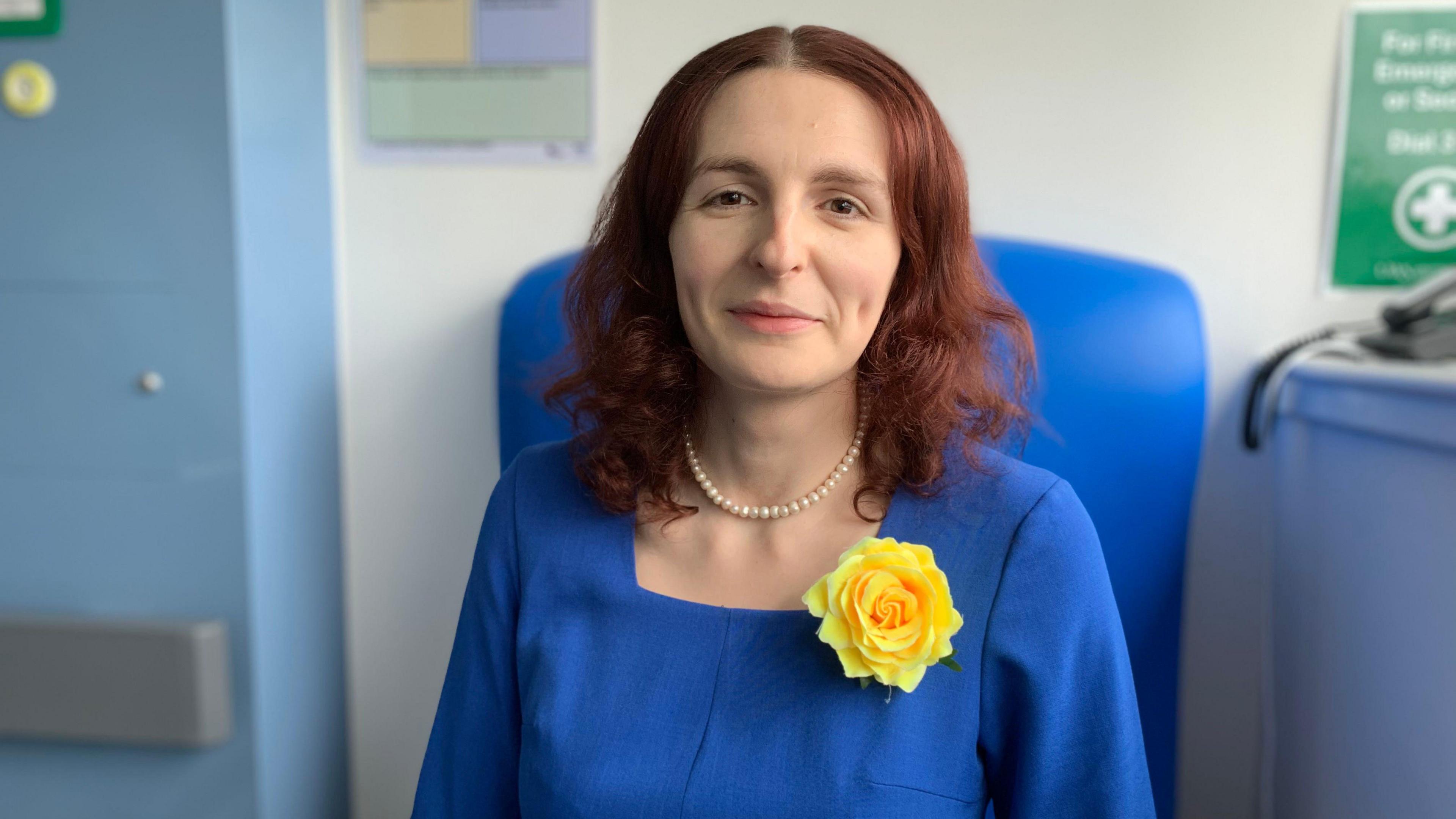A young woman with long reddish-brown hair in a blue dress with a large yellow flower and a pearl-like necklace. She is sitting on a blue chair.