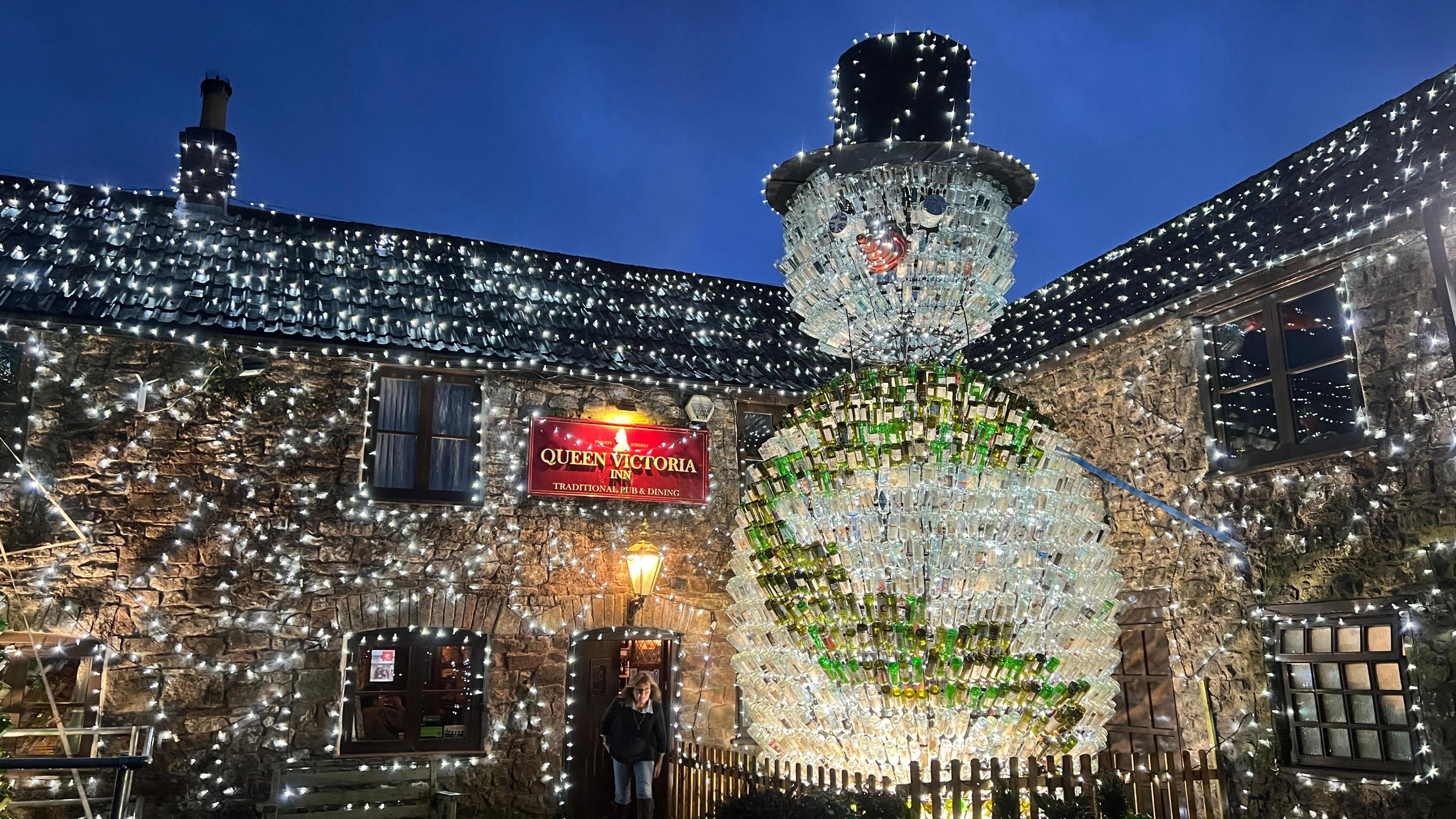 An illuminated snowman covered in wine bottles with pretty bright white lights on the outside of the pub 