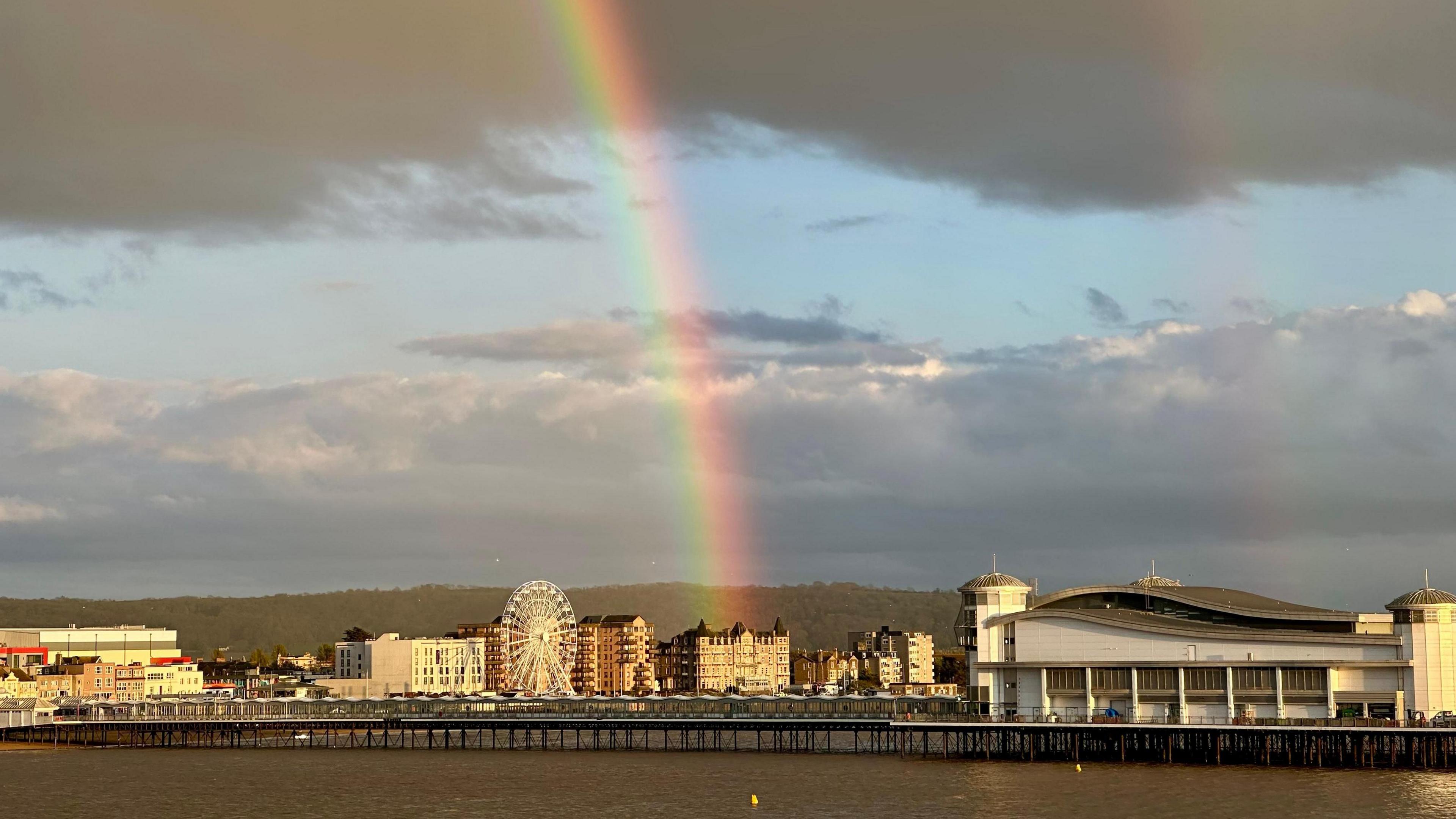 A rainbow appears in a partly cloudy sky with a pier over the sea in the foreground.
