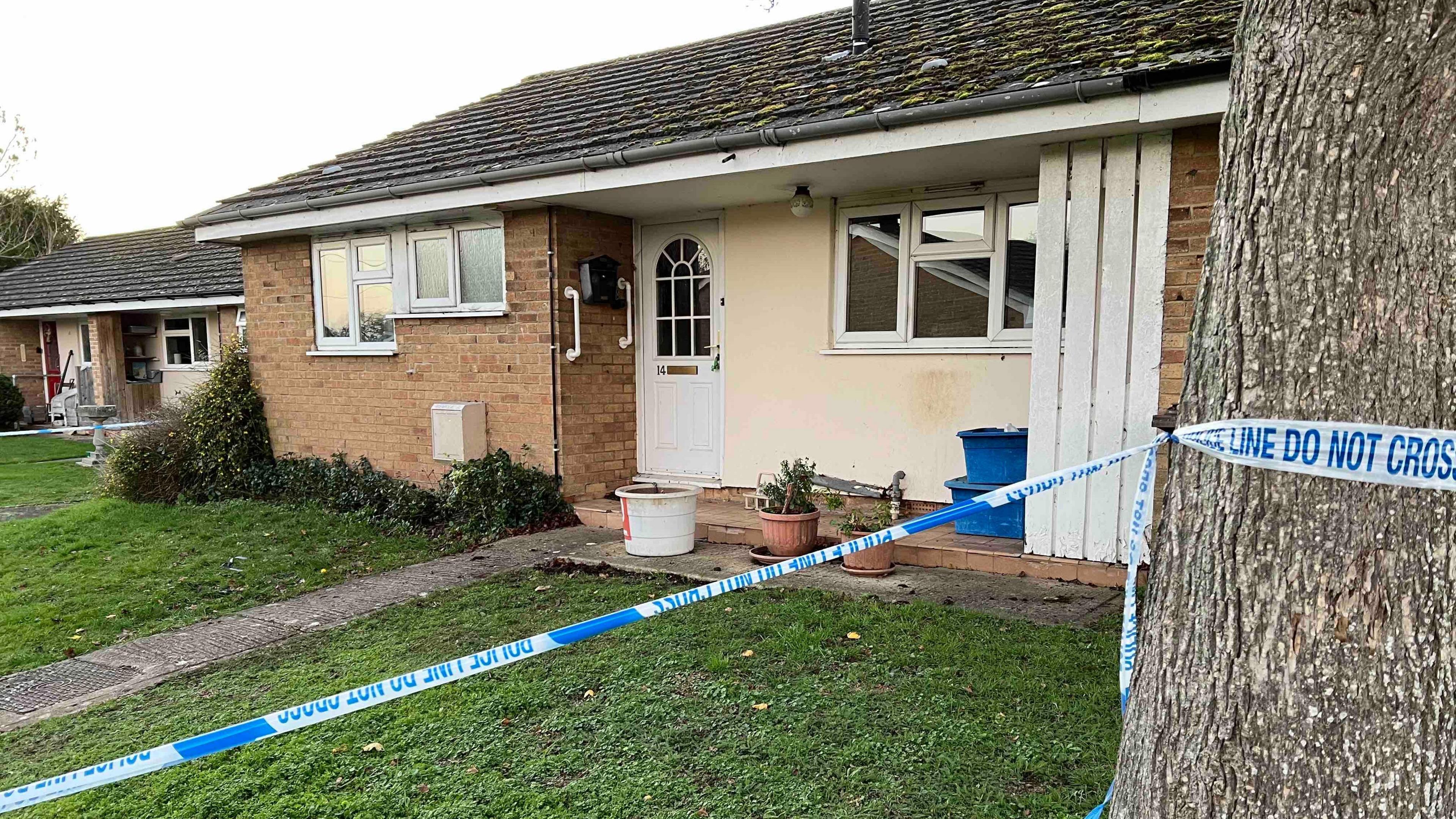A police cordon around a red-brick house with a white door, a cream wall red brick. There are some pot plants and a bucket outside.