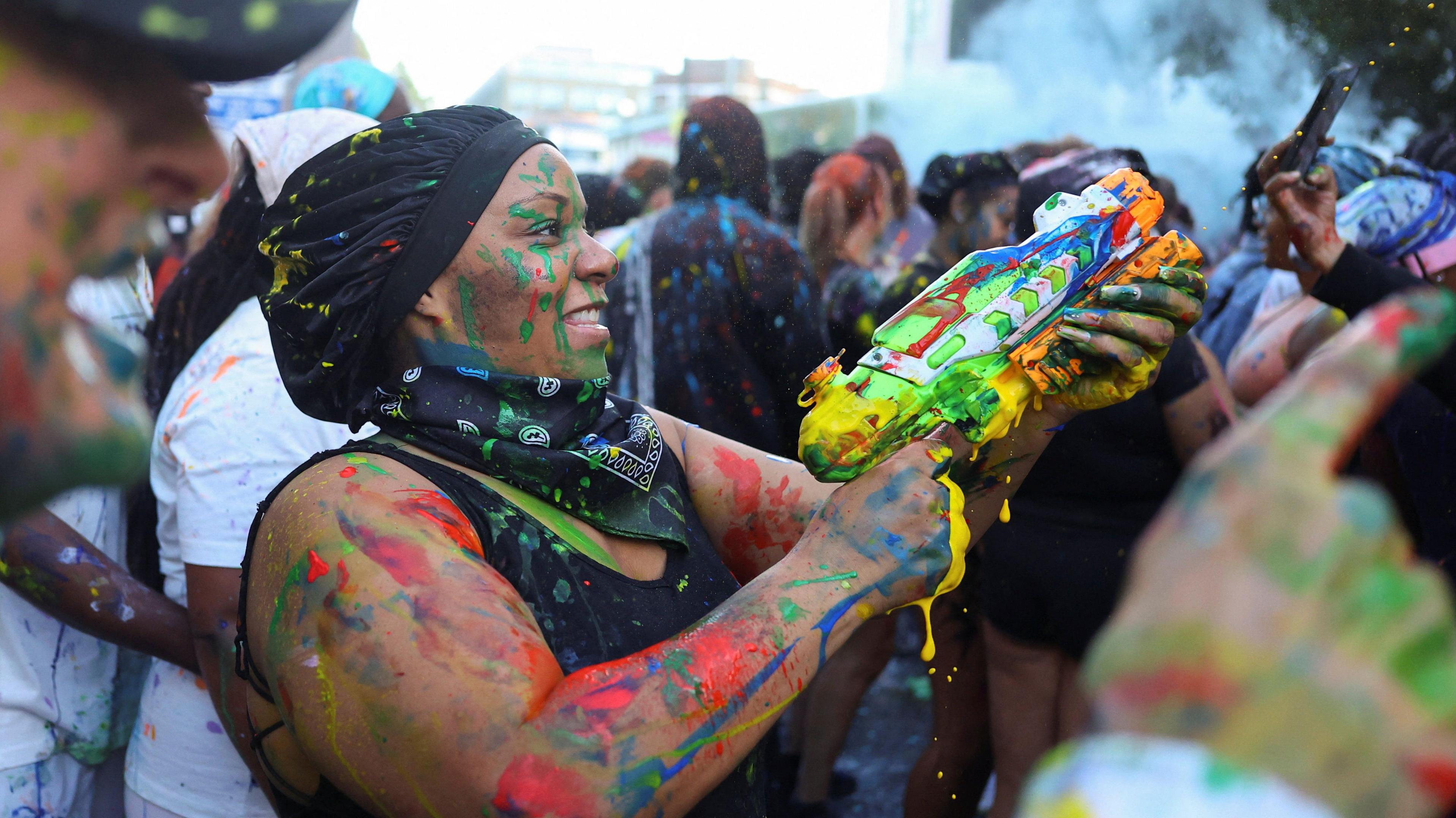 A woman covered in paint holds a water gun filled with paint and points it into a crowd of people. 