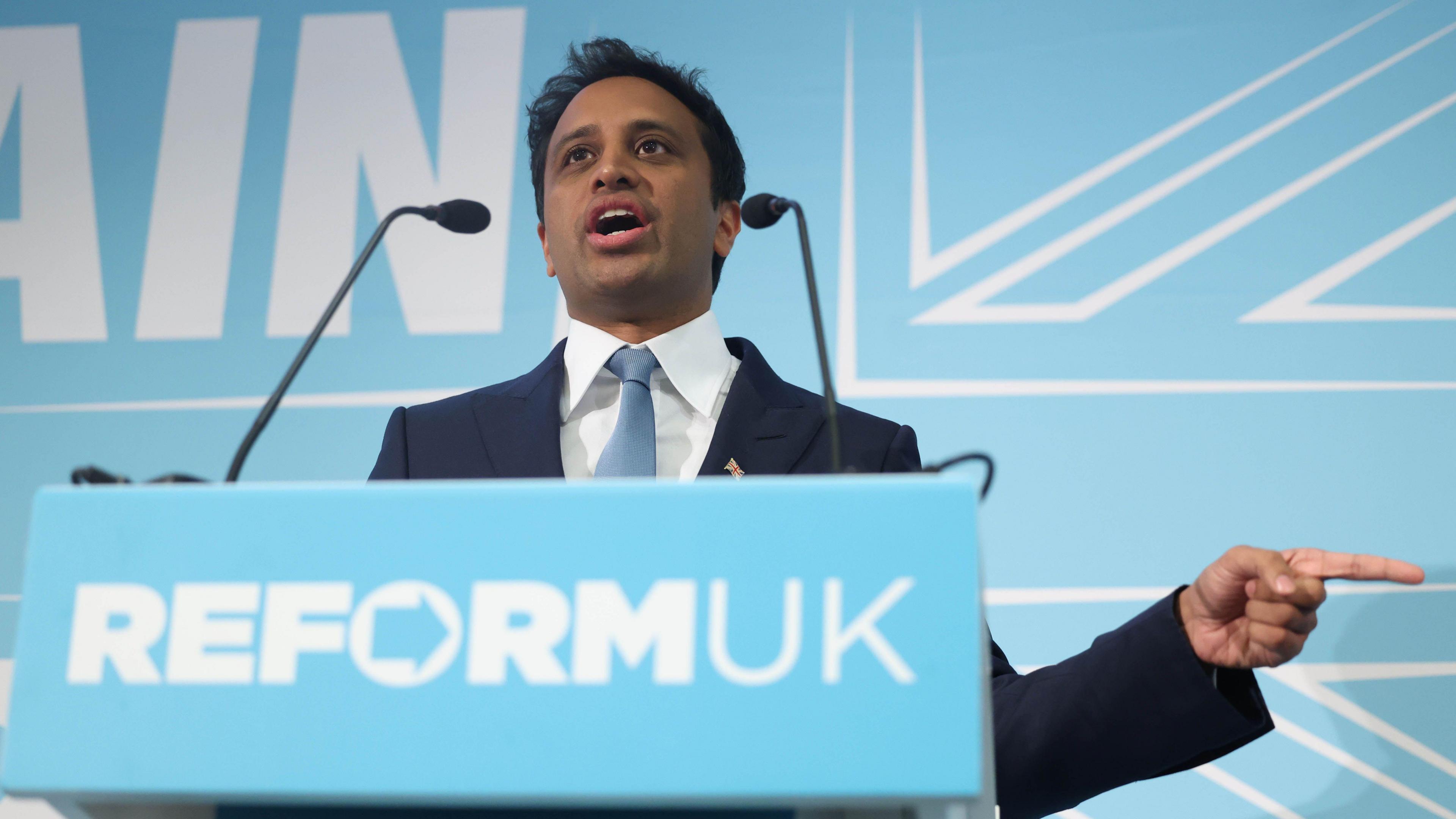 Zia Yusuf, stands at a podium with Reform UK's logo on the front. He has short brown hair and is wearing a suit with a light blue tie.