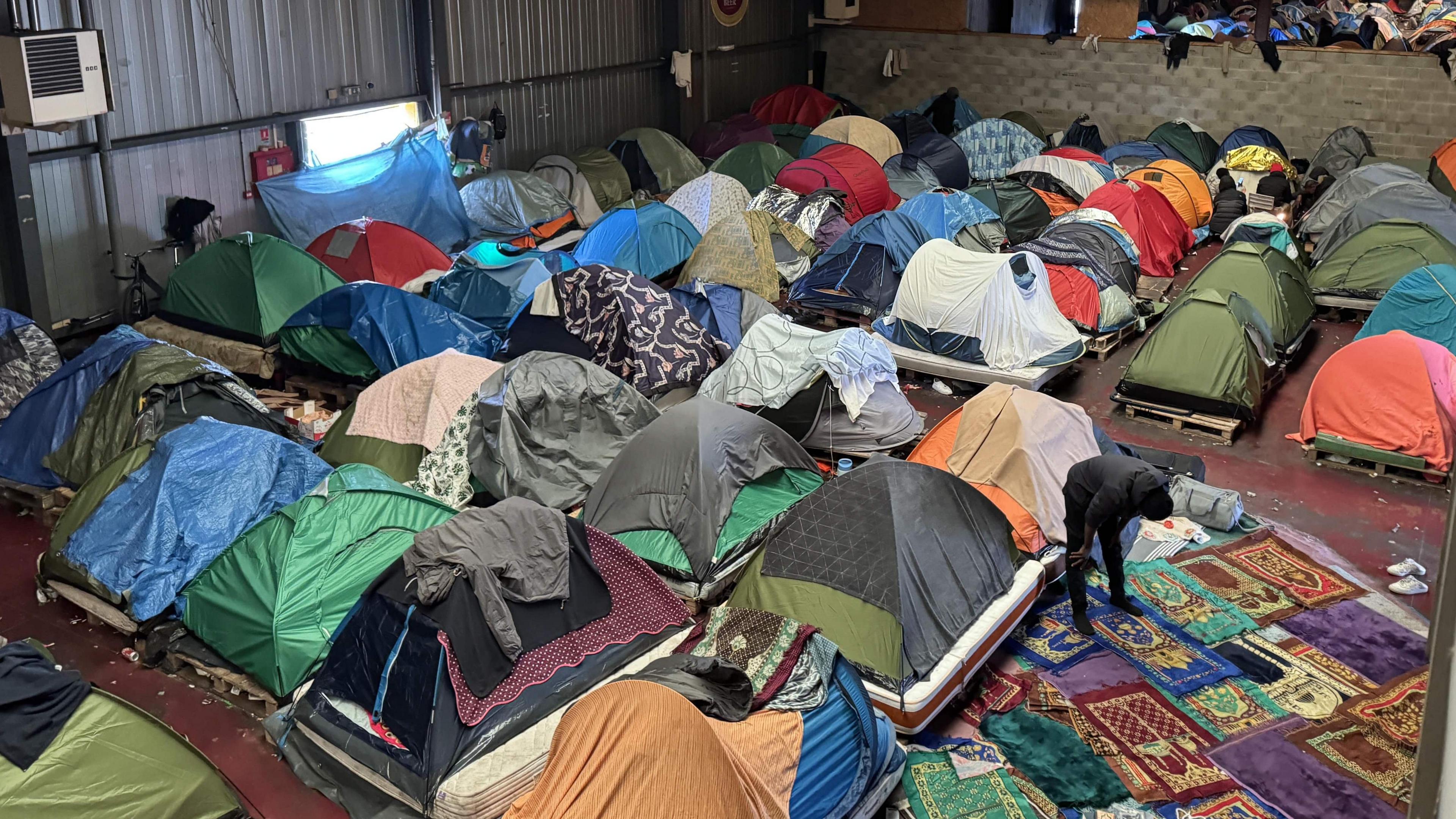 Several tents inside a warehouse. Prayer mats form a makeshift area for those seeking to pray. 