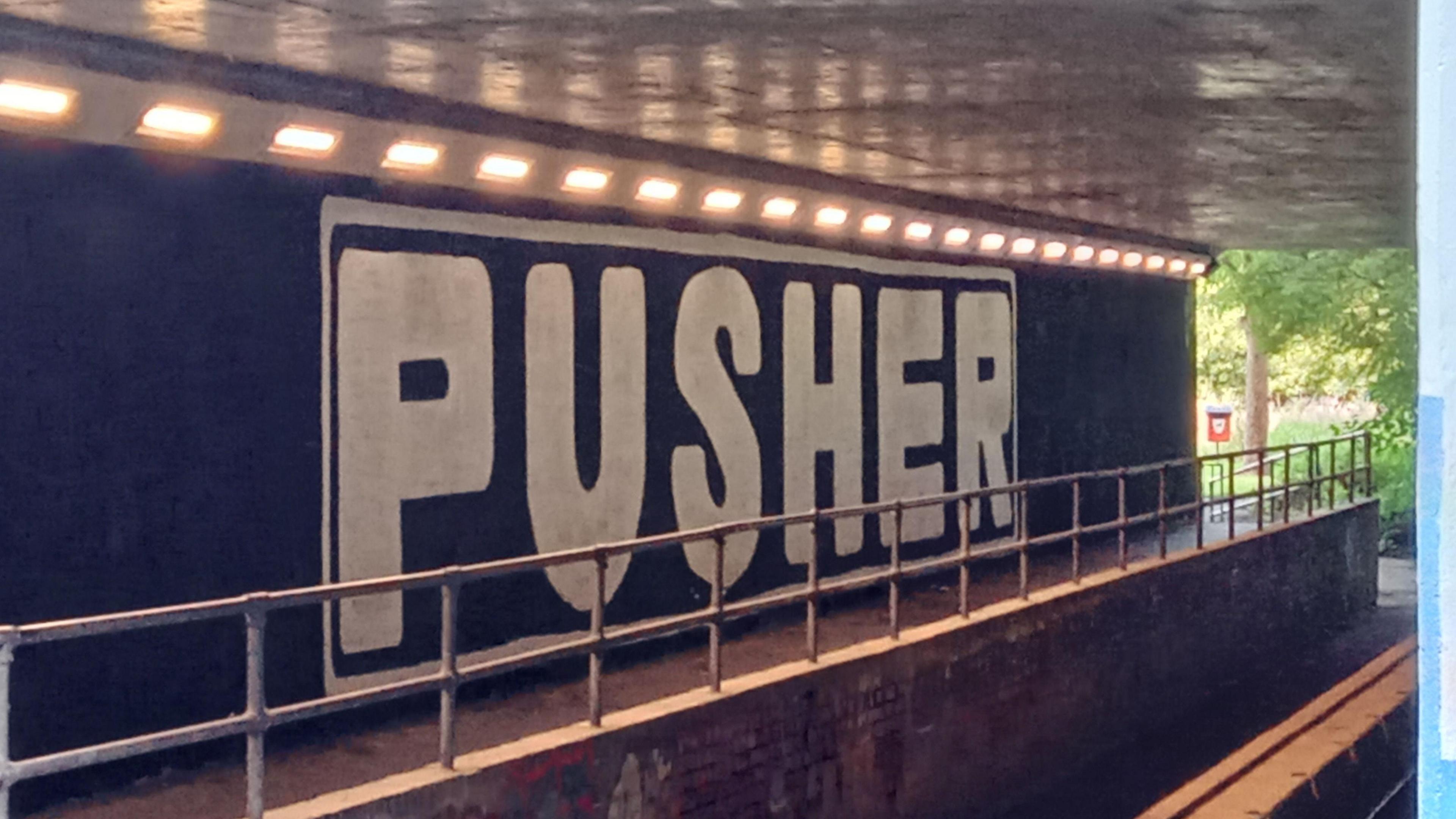 Black paint covering large underpass wall with the words "pusher" written across the area in white.