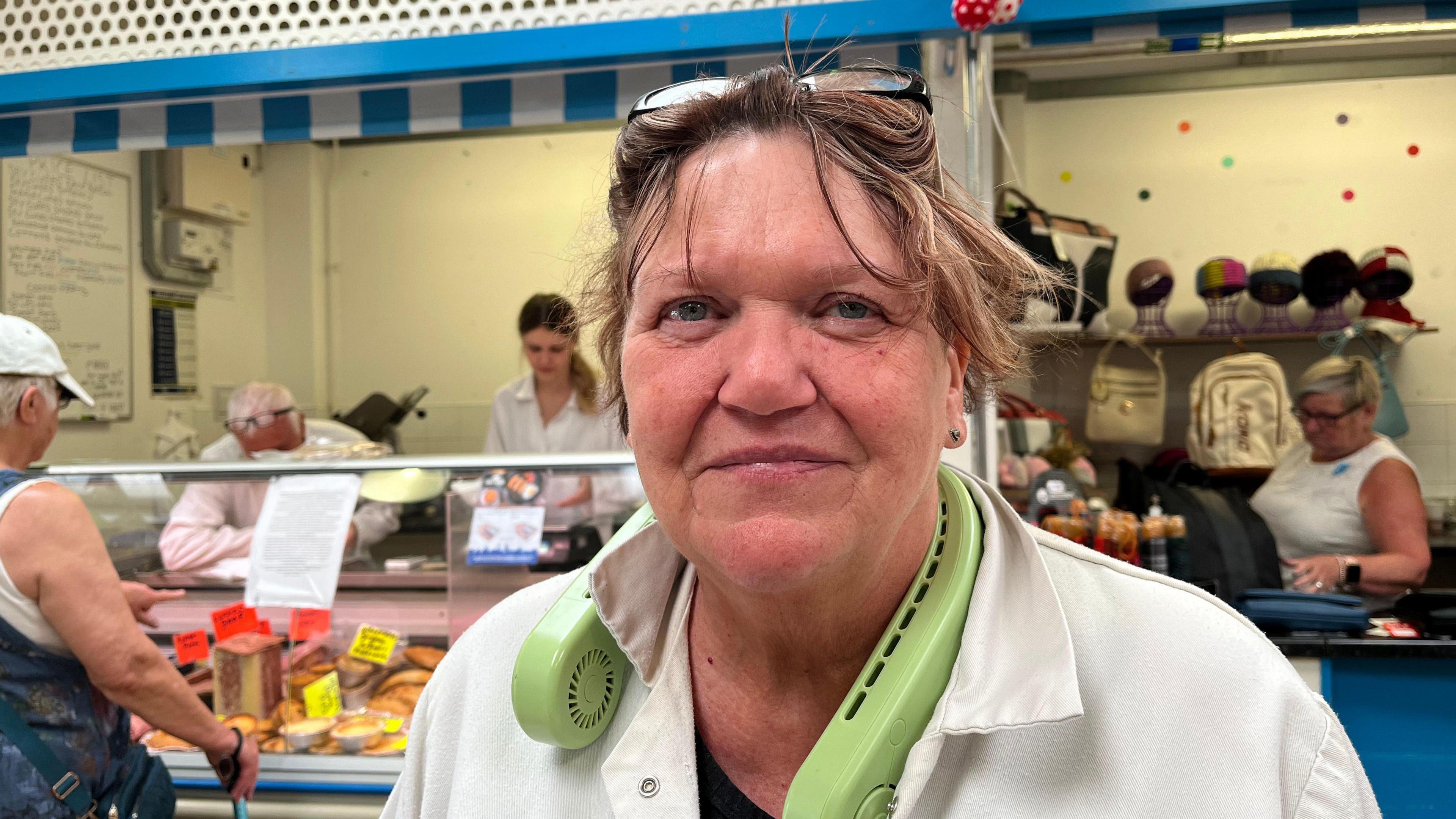 Mary Walley stands smiling in front of a butcher's stall in a white top, with a light green neck fan around her neck, with her hair tied up and glasses on her head.