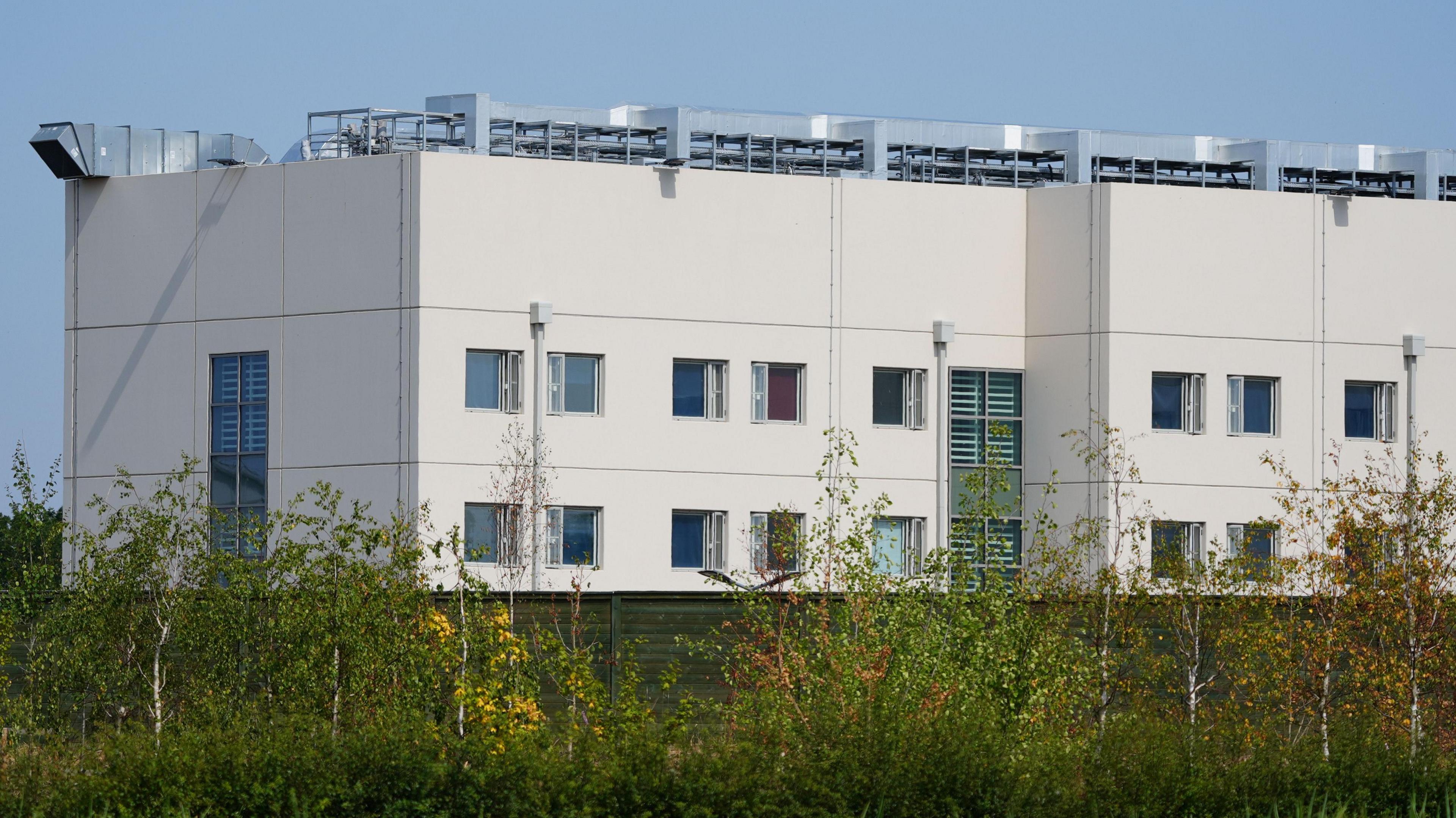 A modern-looking, white prison building. It appears to be four stories high with the central stories having two rows of windows. A ventilation system can be seen on the roof. Trees are growing in the foreground.