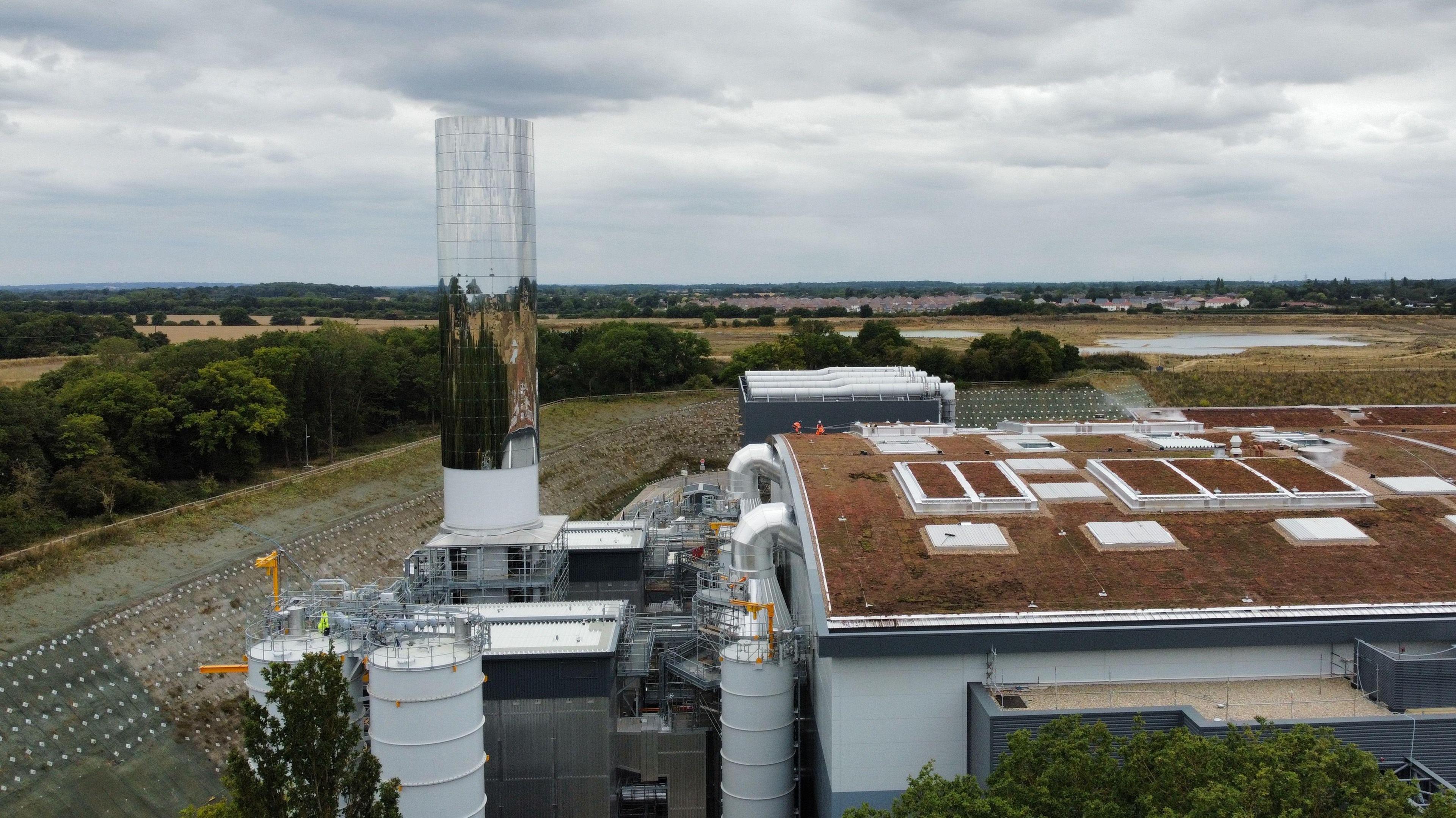 The Integrated Waste Management Facility (IWMF) at Rivenhall, which is an incinerator designed to burn Essex County Council's household waste. A drone photograph shows the domed roof of the incinerator building, covered with browned grass. There are several white hatches in the roof of the building. Next to it are several large pipes, and a 35-metre tall chimney which is covered in a reflective surface. There are green fields and trees in the distance.