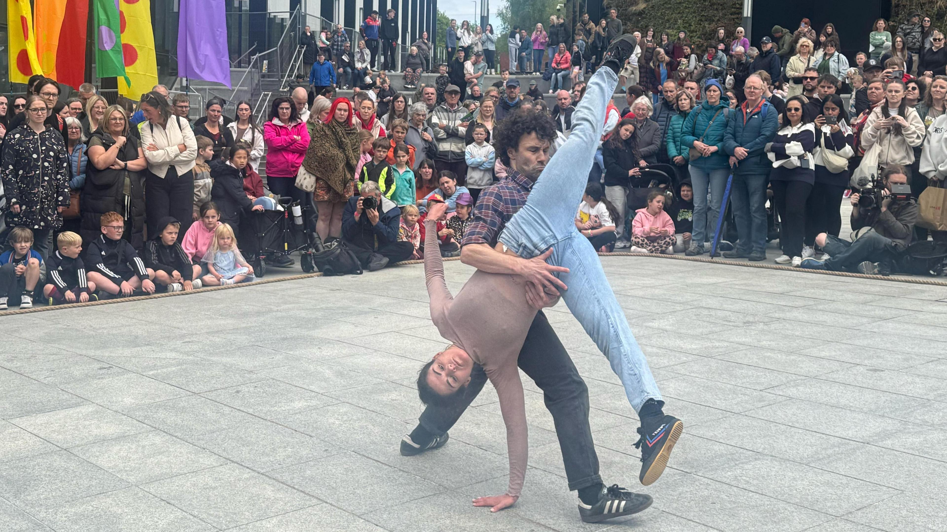 Male dancer twirls woman around in a cartwheel with crowds watching behind and various colourful banners fluttering behind.