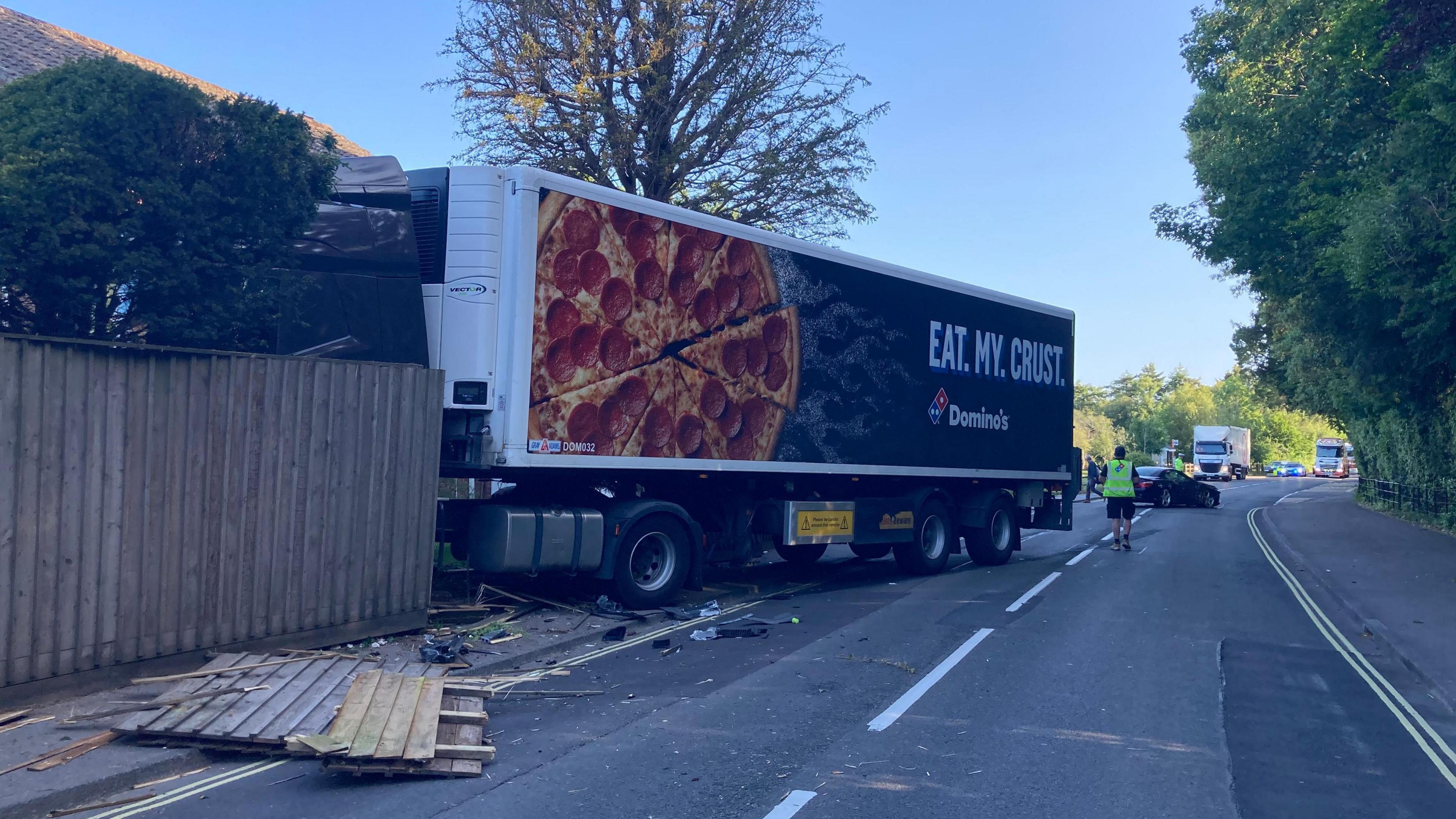 Side of lorry protruding from a garden fence with debris across the road a man in high-vis jacket is in the middle of the road with a car across and other vehicles queueing behind.