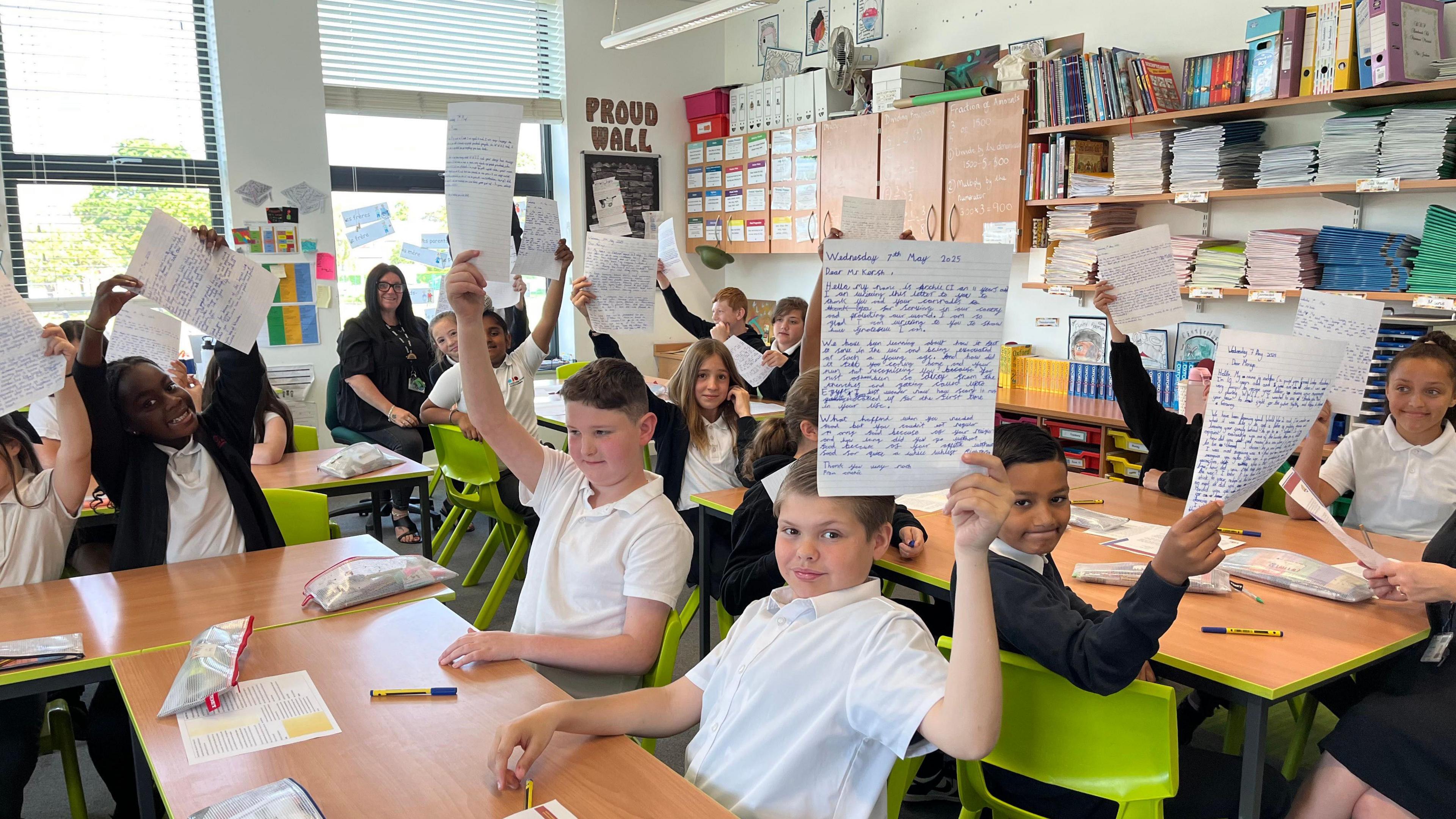 Children hold up the letters they've written, whilst sitting in their classroom