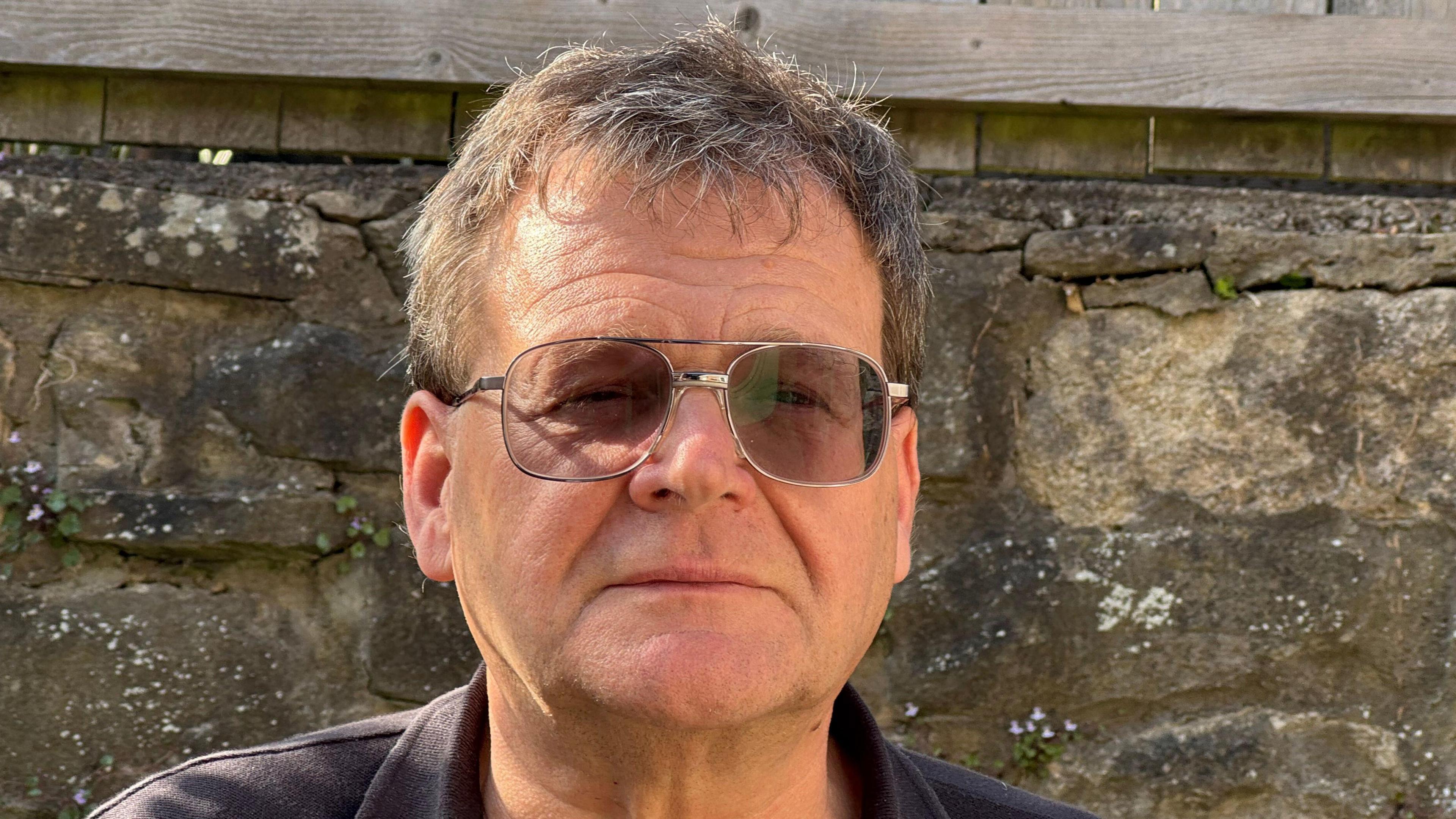 Professor Paul Christensen looks at the camera while standing in front of a stone wall. He has short dark hair with some grey in it. He wears glasses and a blue polo top.