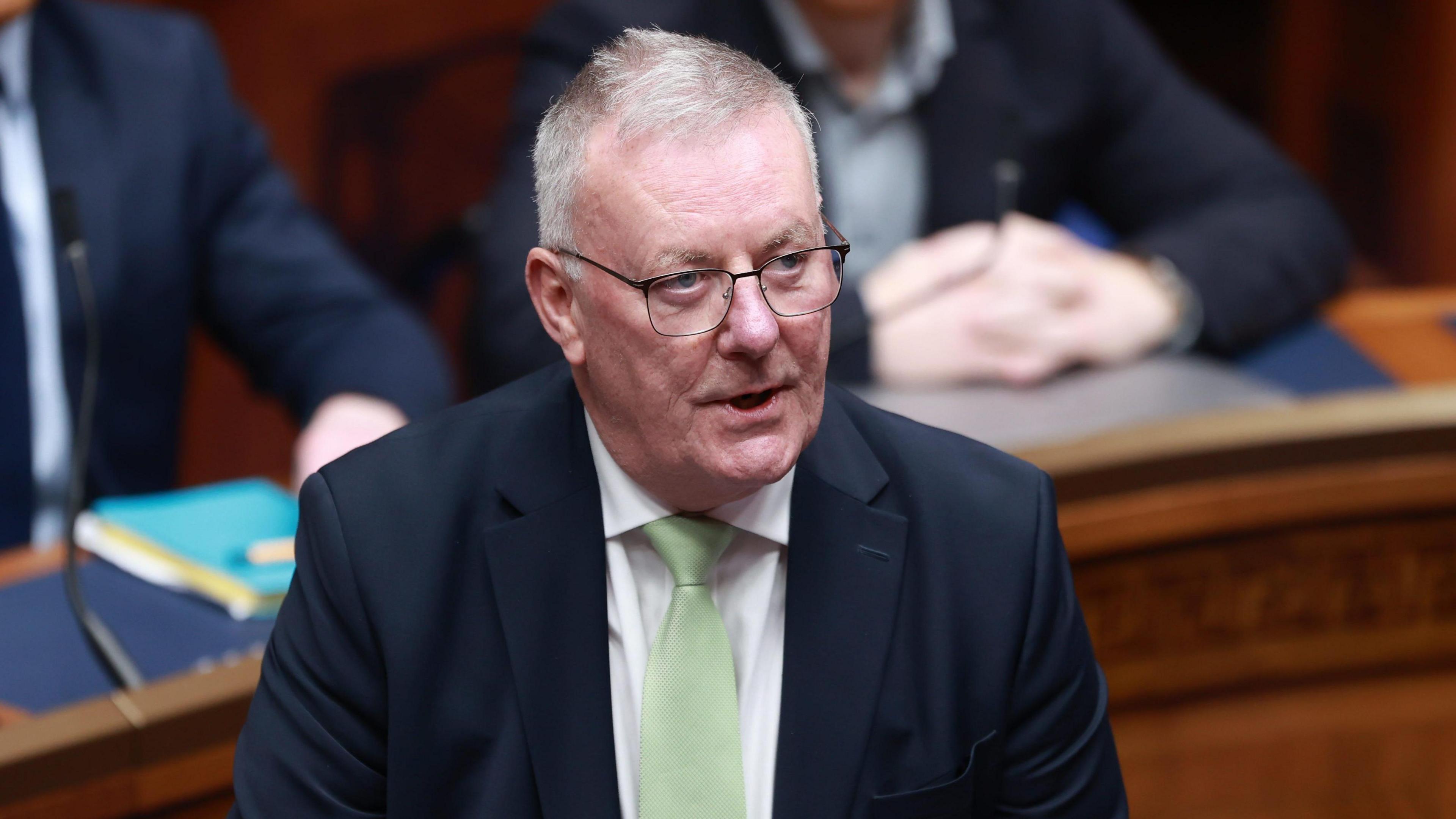 Mike Nesbitt, he has grey hair and is wearing glasses. He is wearing a navy suit with light green tie