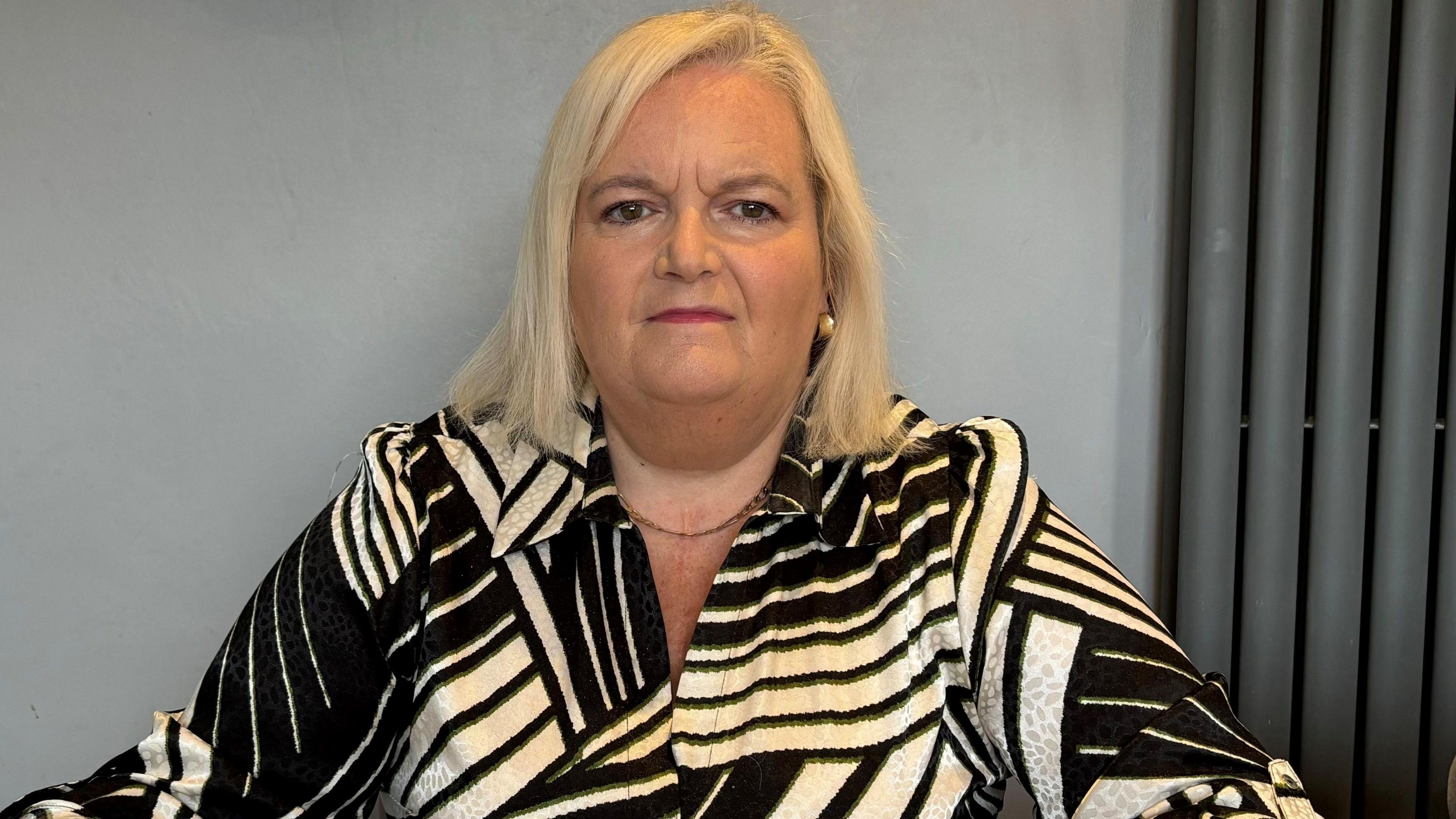 A woman is looking directly into the camera. She has blonde, shoulder-length hair and is wearing a black and cream patterned shirt. The wall behind her is grey and to the side there is a horizontal dark grey radiator.