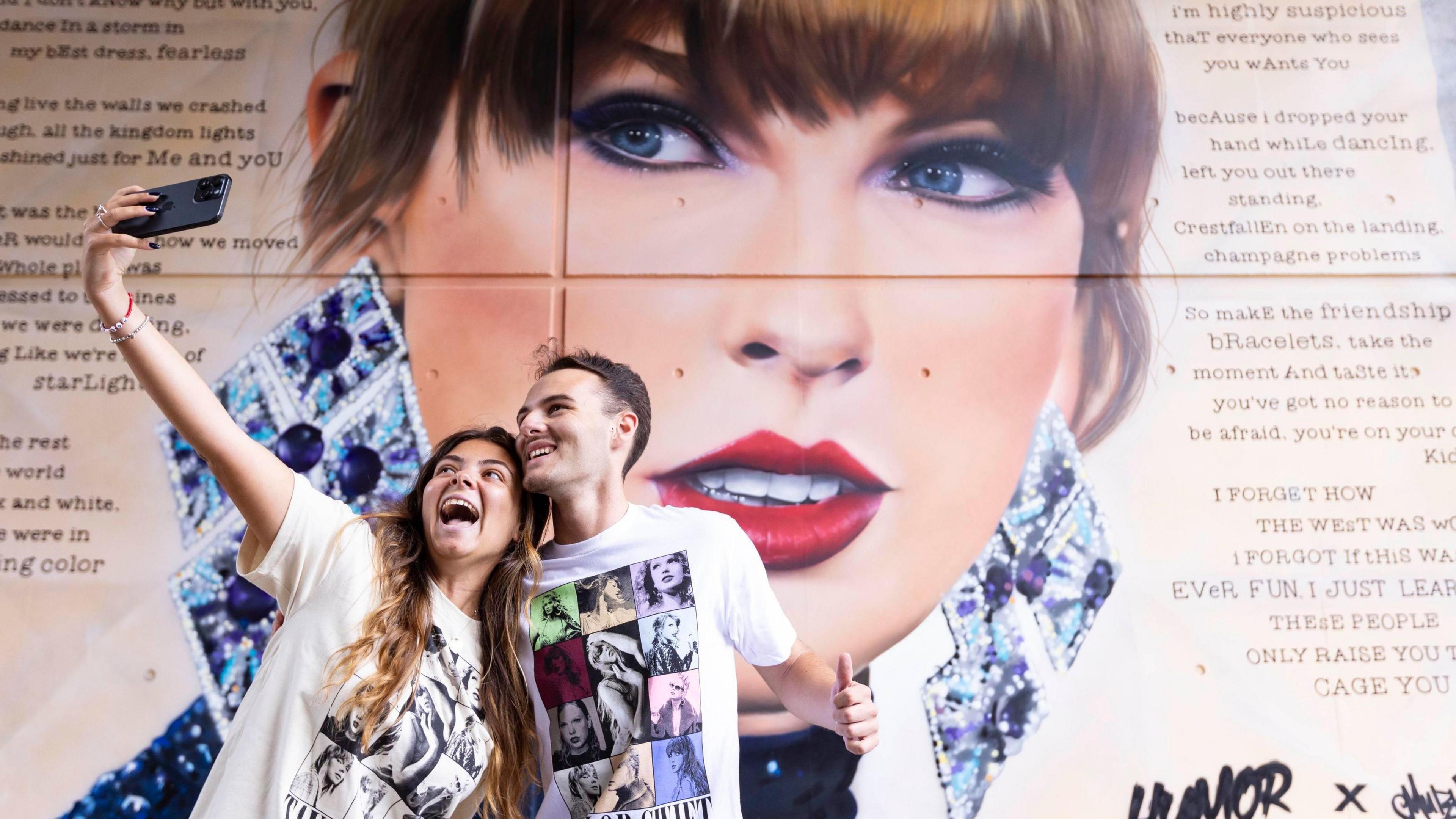 Two people take a selfie in front of a Taylor Swift mural