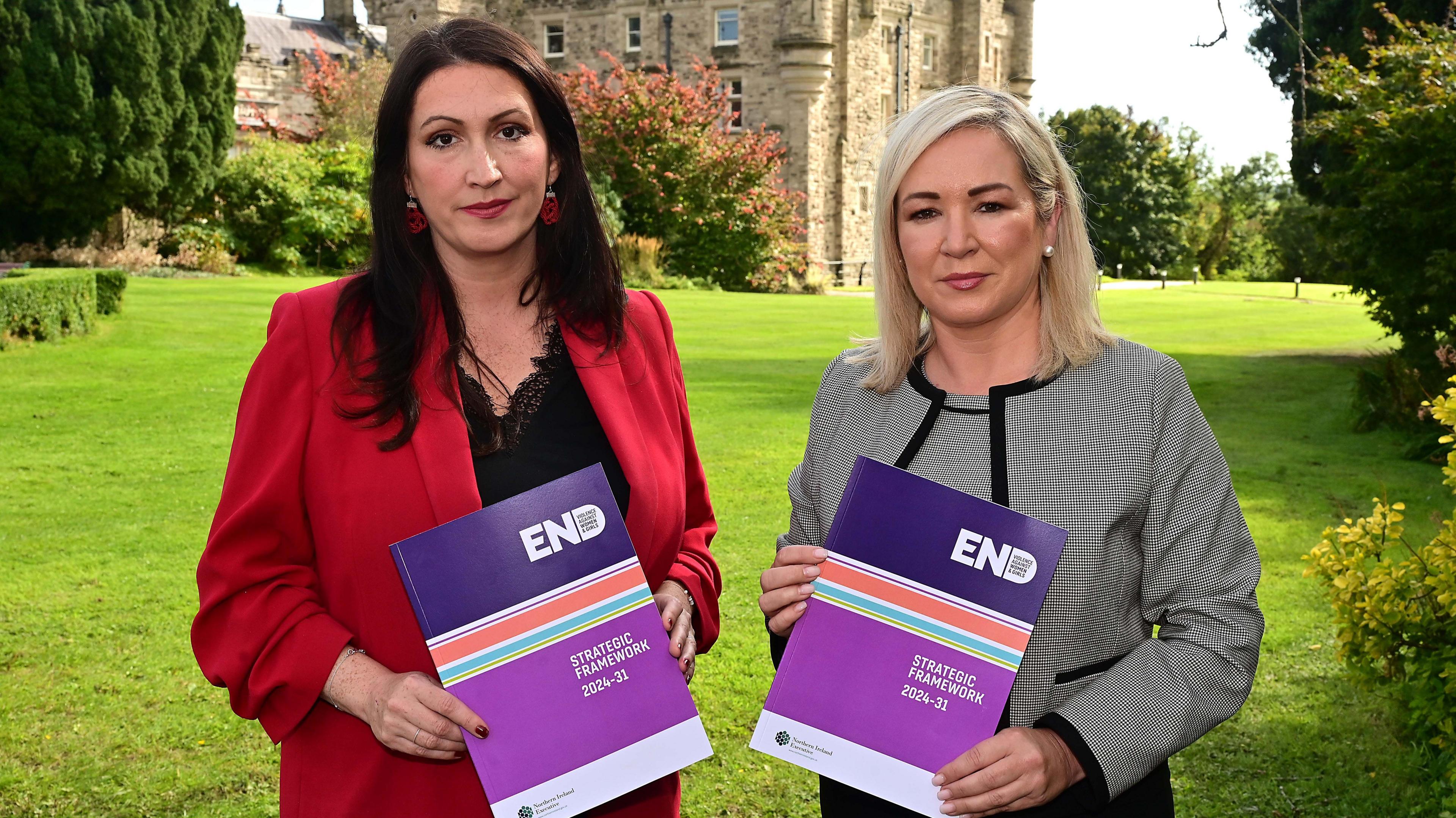 Deputy First Minister Emma Little-Pengelly in a red suit and First Minister Michelle O'Neill in a grey suit, launched the Ending Violence against Women and Girls strategic framework last year, they are holding the paperwork on the lawn at Stormont estate with castle in background