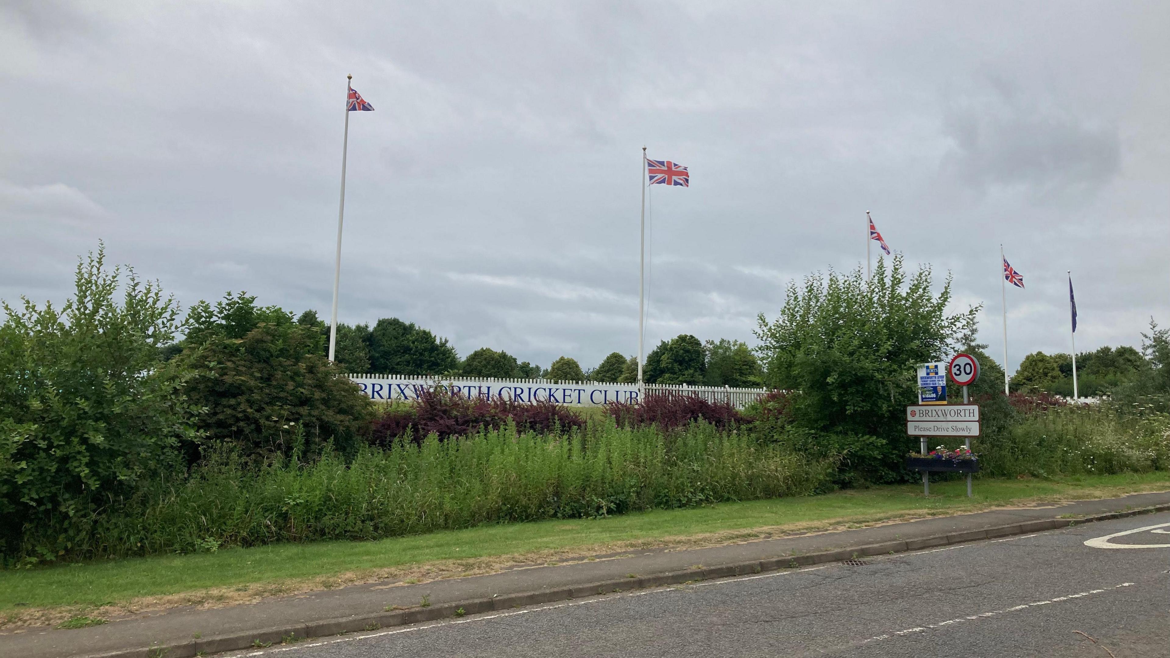 In the foreground of the picture is a road. The other side of the road a sign that says "Brixworth Cricket Club" is just visible behind an overgrown hedge. Union flags on tall flag poles are flying in front of a cloudy sky. 