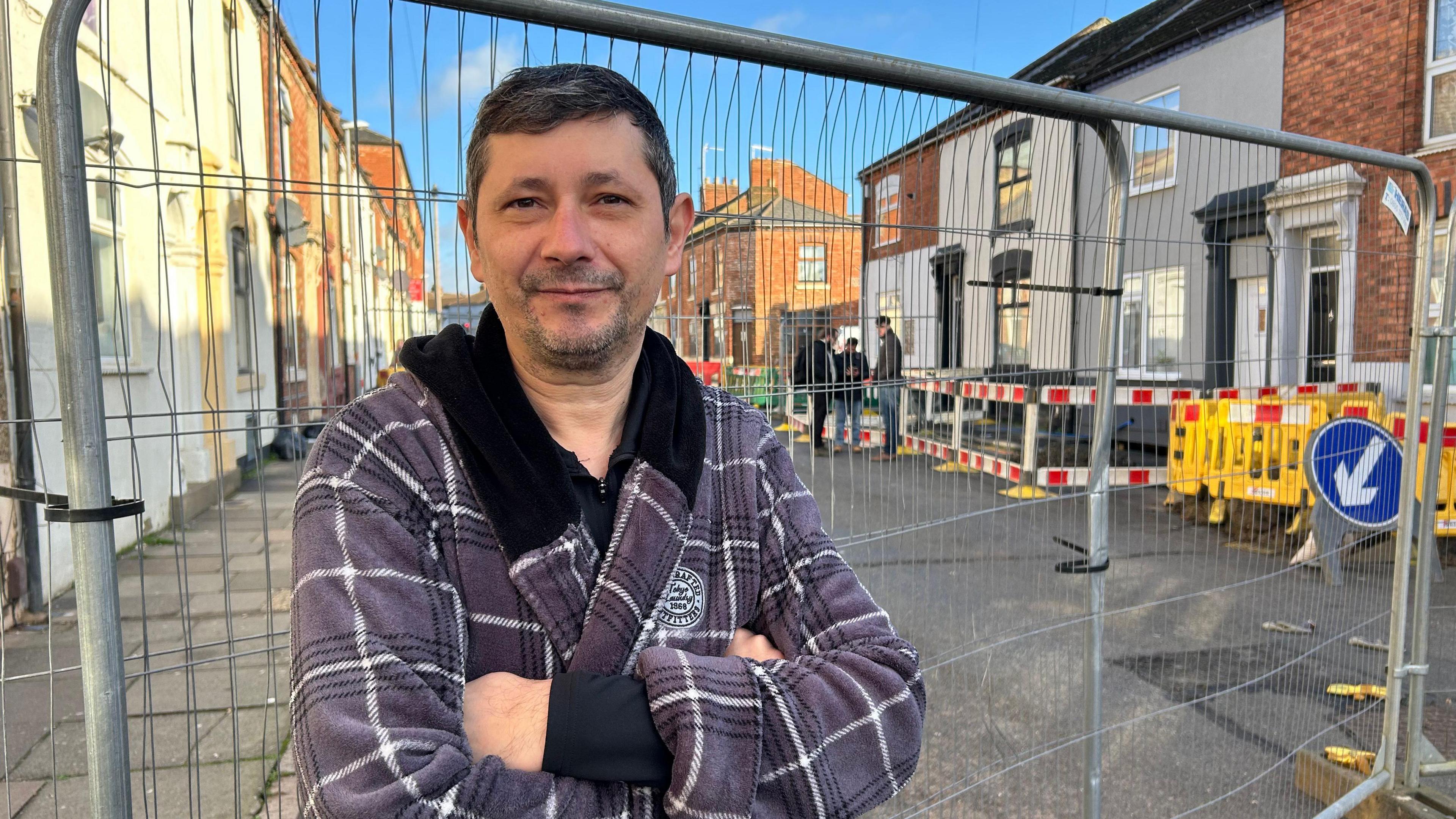 A man in a purple grey chequered dressing gown stands by a metal fence in the middle of a terraced street. Behind him are three people discussing the sink hole which is hidden behind red and white barriers. 