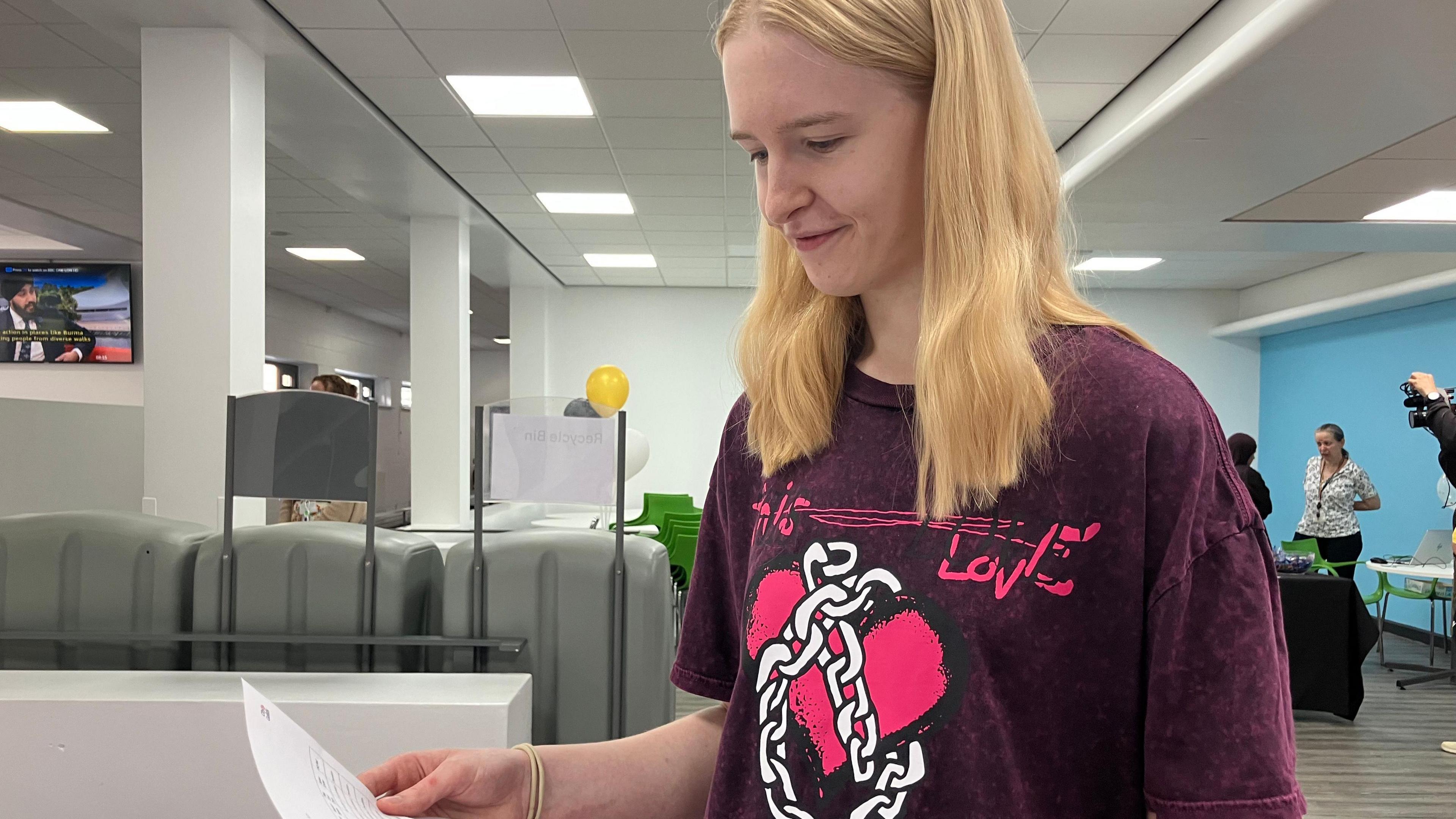 Emily Griffin looks down at a piece of paper with her results on. She has blond mid-length hair styled in a centre parting and wears a burgundy short sleeved top which has an image of a cartoon heart wrapped in a chain. In the background are some desks and people talking.