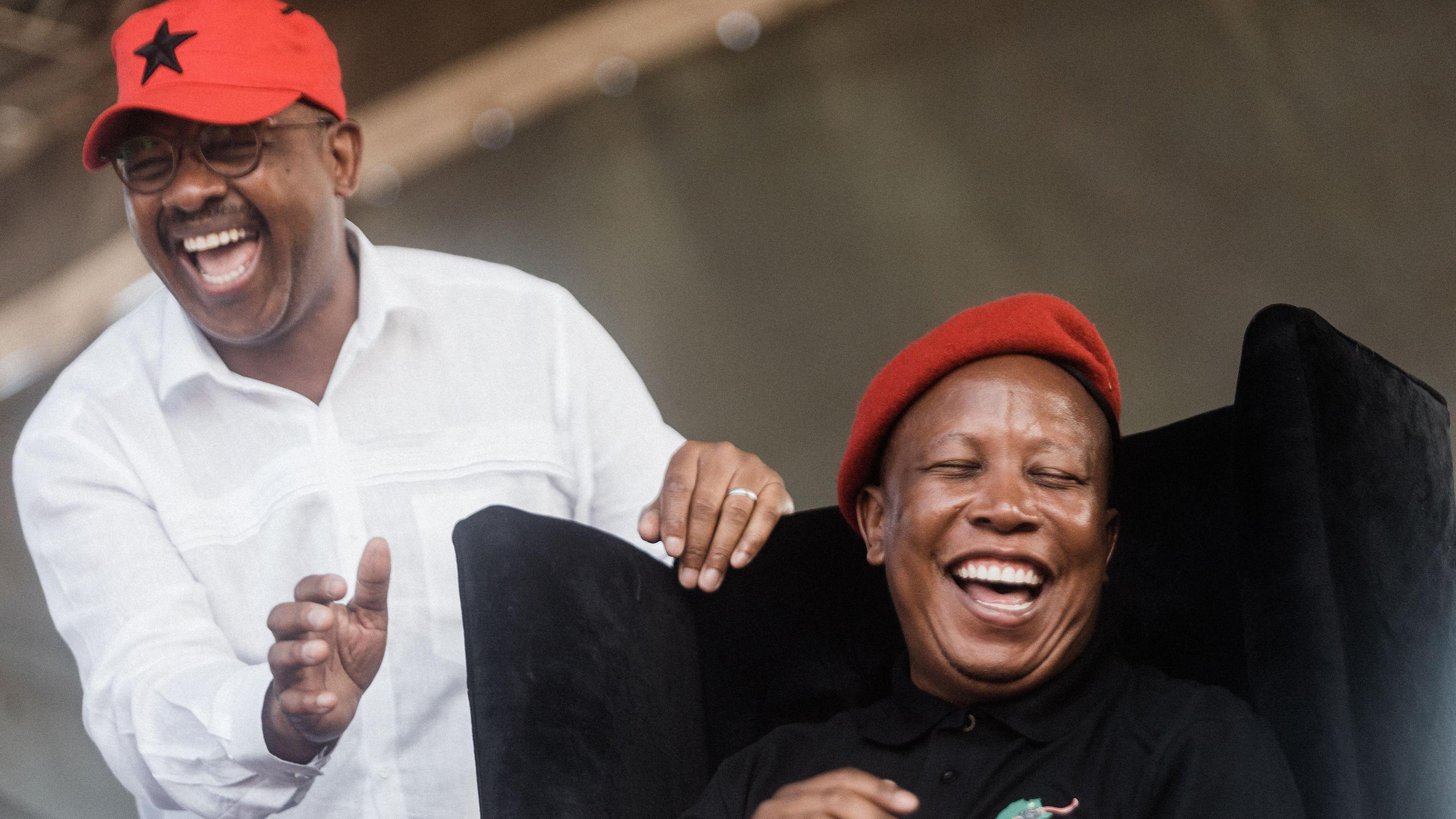 Two men in the picture. One man on the left, Dali Mpofu, wearing a white shirt and a red cap. The man on the right, Julius Malema, is wearing a red cap and a black top.