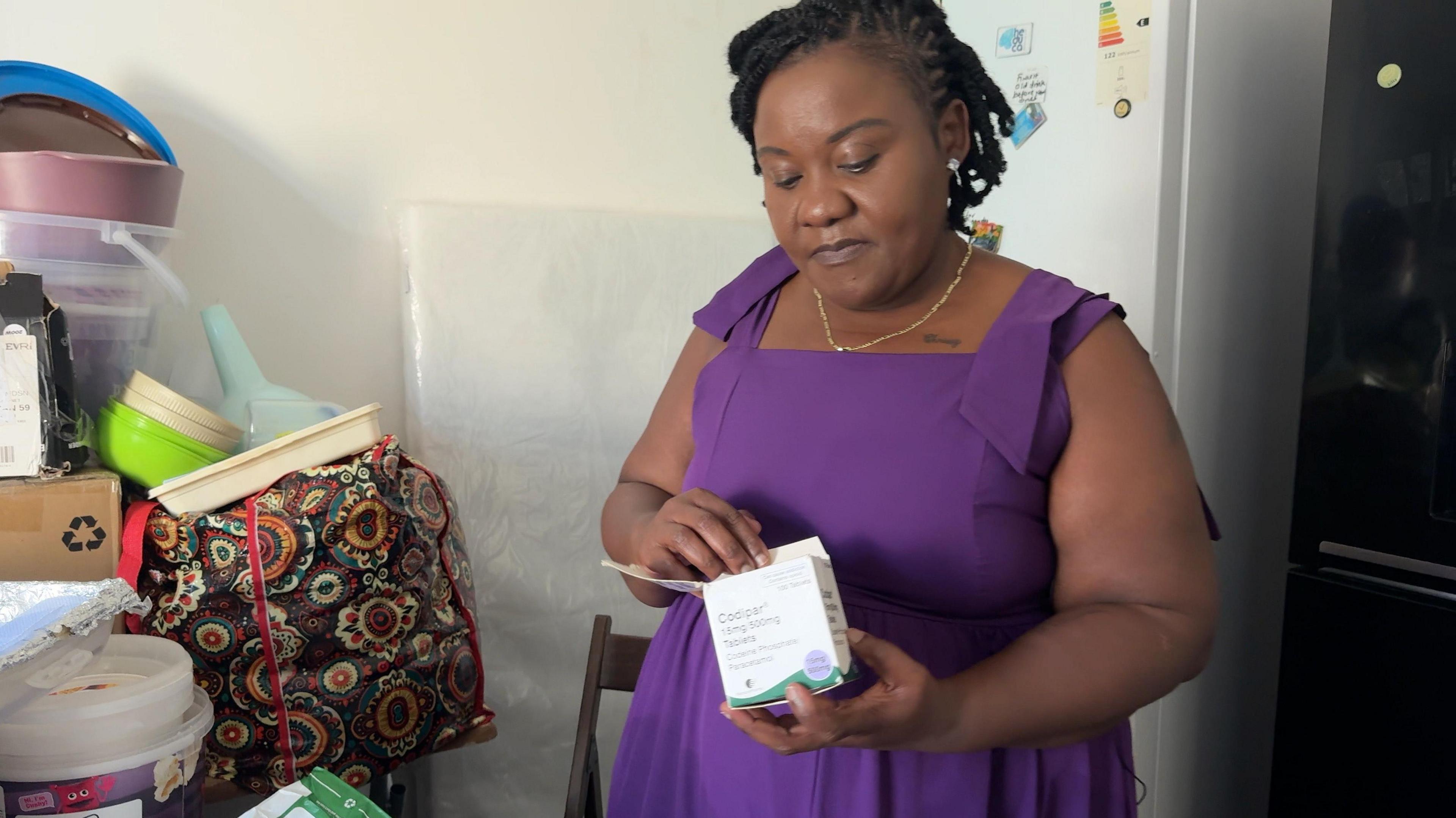 Christiana is looking at a box of tablets as she stands in her kitchen. She is wearing a purple dress and there are bags and items piled up on the left side of the picture.