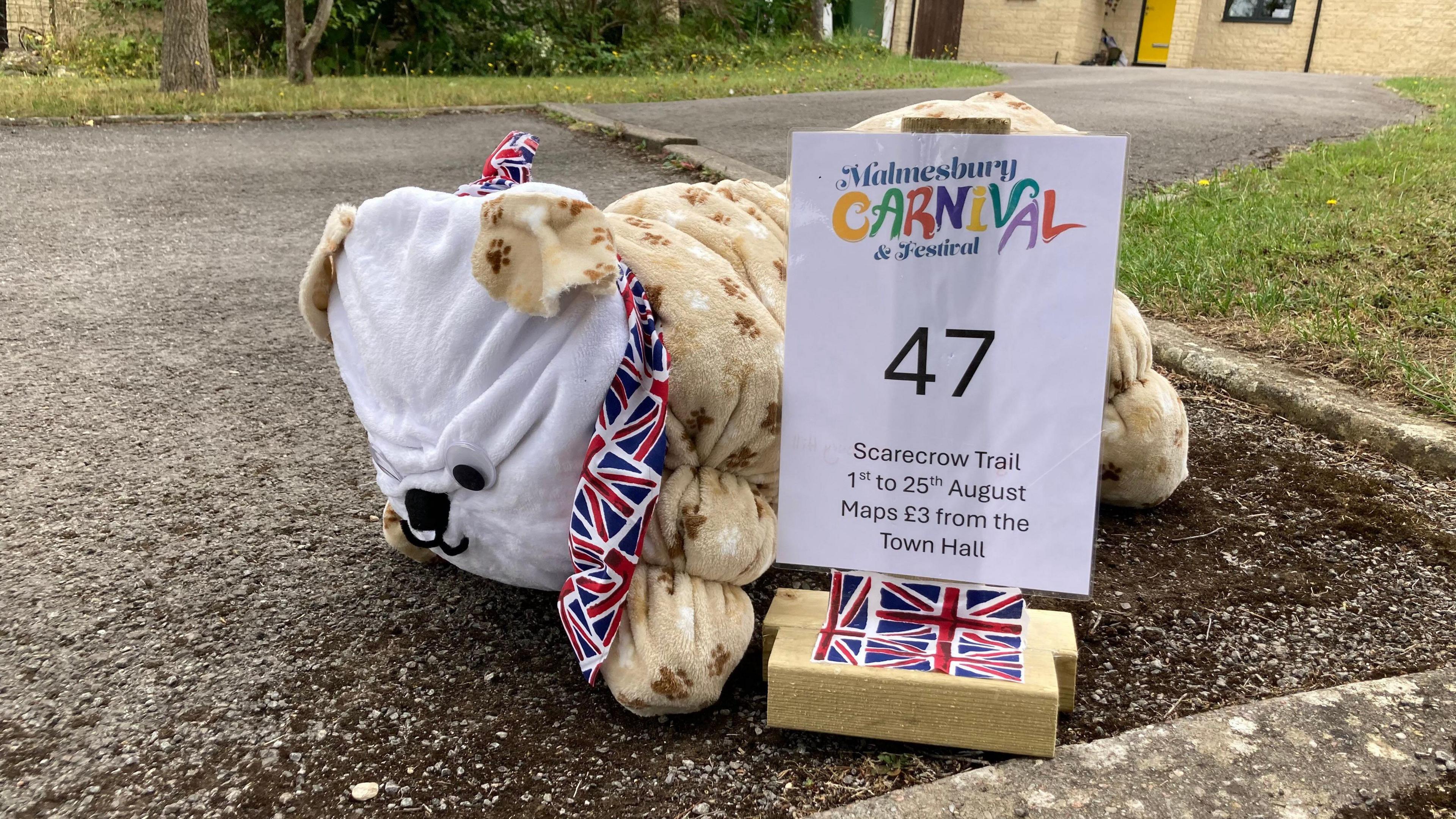 A scarecrow of a British Bulldog. It is white and beige. There is a sign advertising the Malmesbury Carnival and Scarecrow Trail next to it.