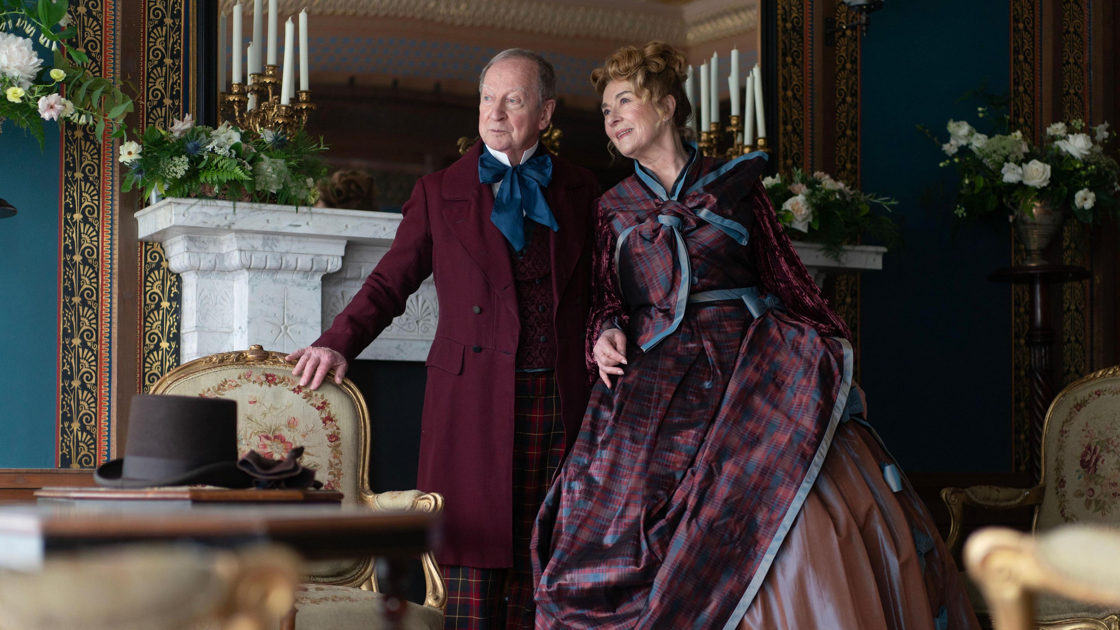 Actor Bill Paterson is dressed in a period costume consisting of a long burgundy jacket, blue cravat and tartan trousers. He is leaning against an old-fashioned chair while standing in front of a white marble fireplace and mirror. Next to him is Julie Cadzow, also dressed in a costume. Her outfit is a burgundy and blue tartan dress and jacket.