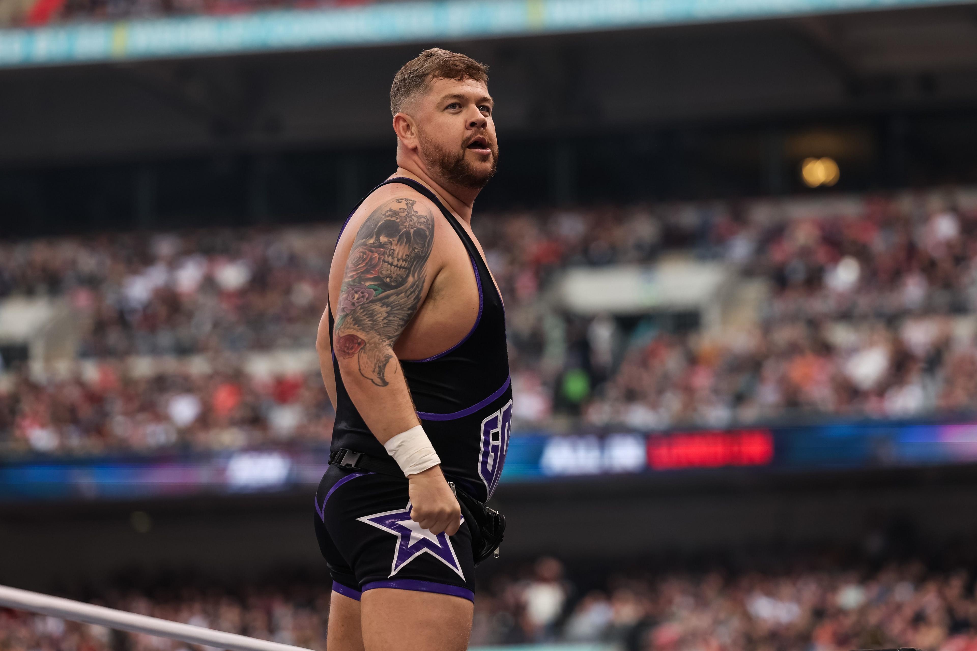 Grado stands side-on in front of crowds wearing a black wrestling leotard - a skull tattoo takes up his entire left shoulder. He has short brown hair and a beard.