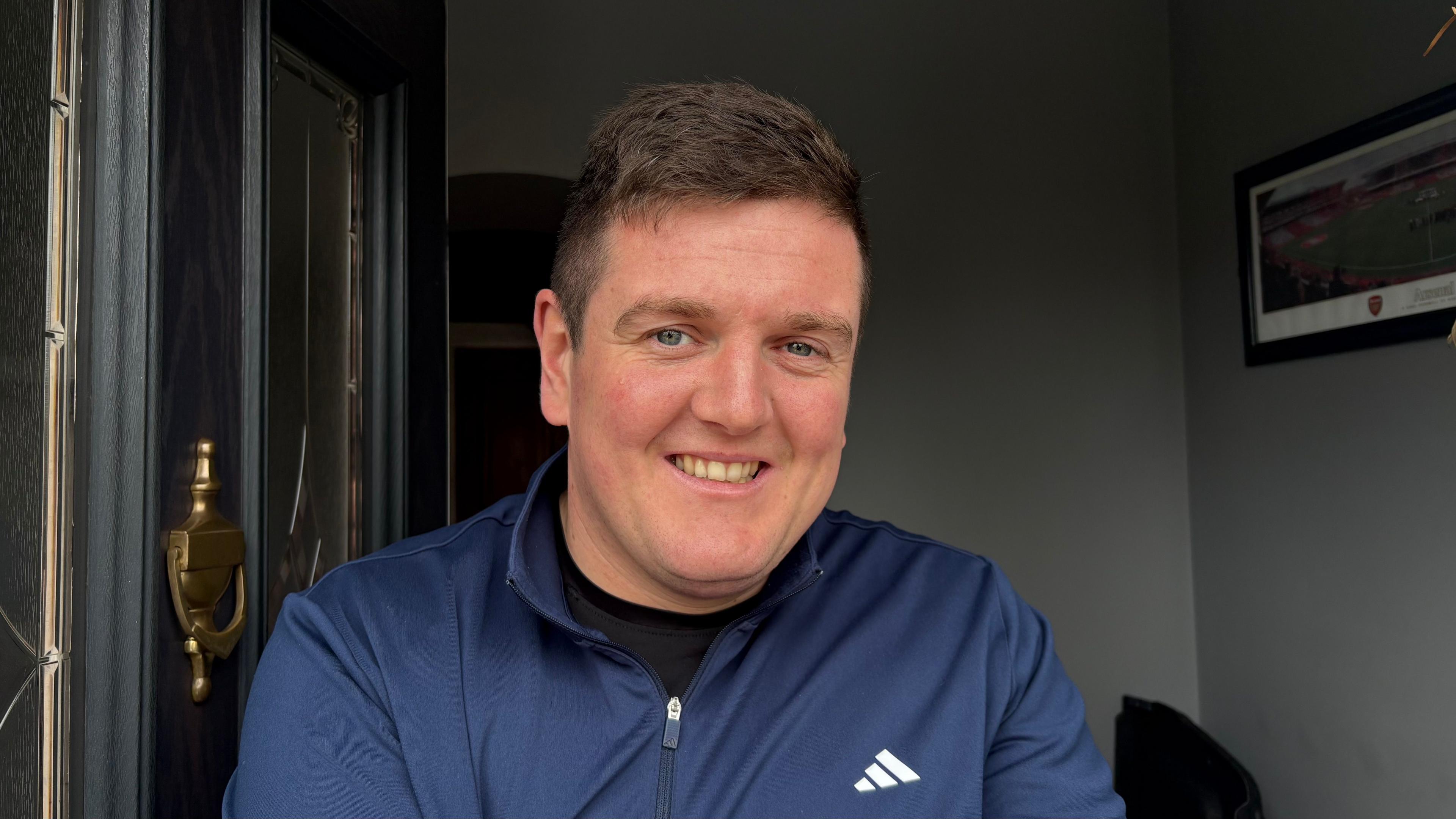 Nick Henderson with short dark hair wearing a black zip-up top with a white logo. He is standing at the front door of his house.