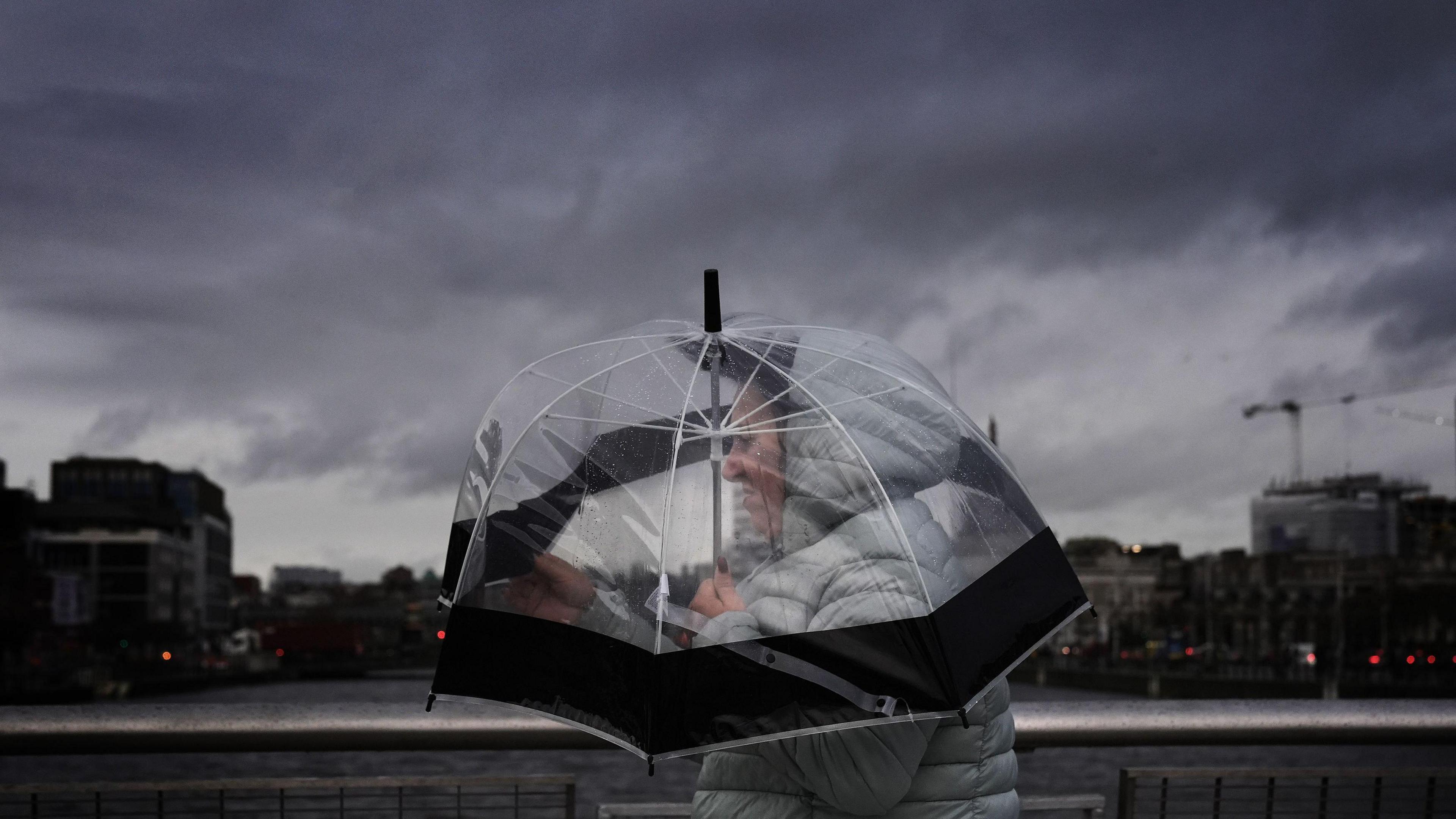 Dark storm clouds over Dublin. Someone wearing a winter coat with their hood up clutches their umbrella.