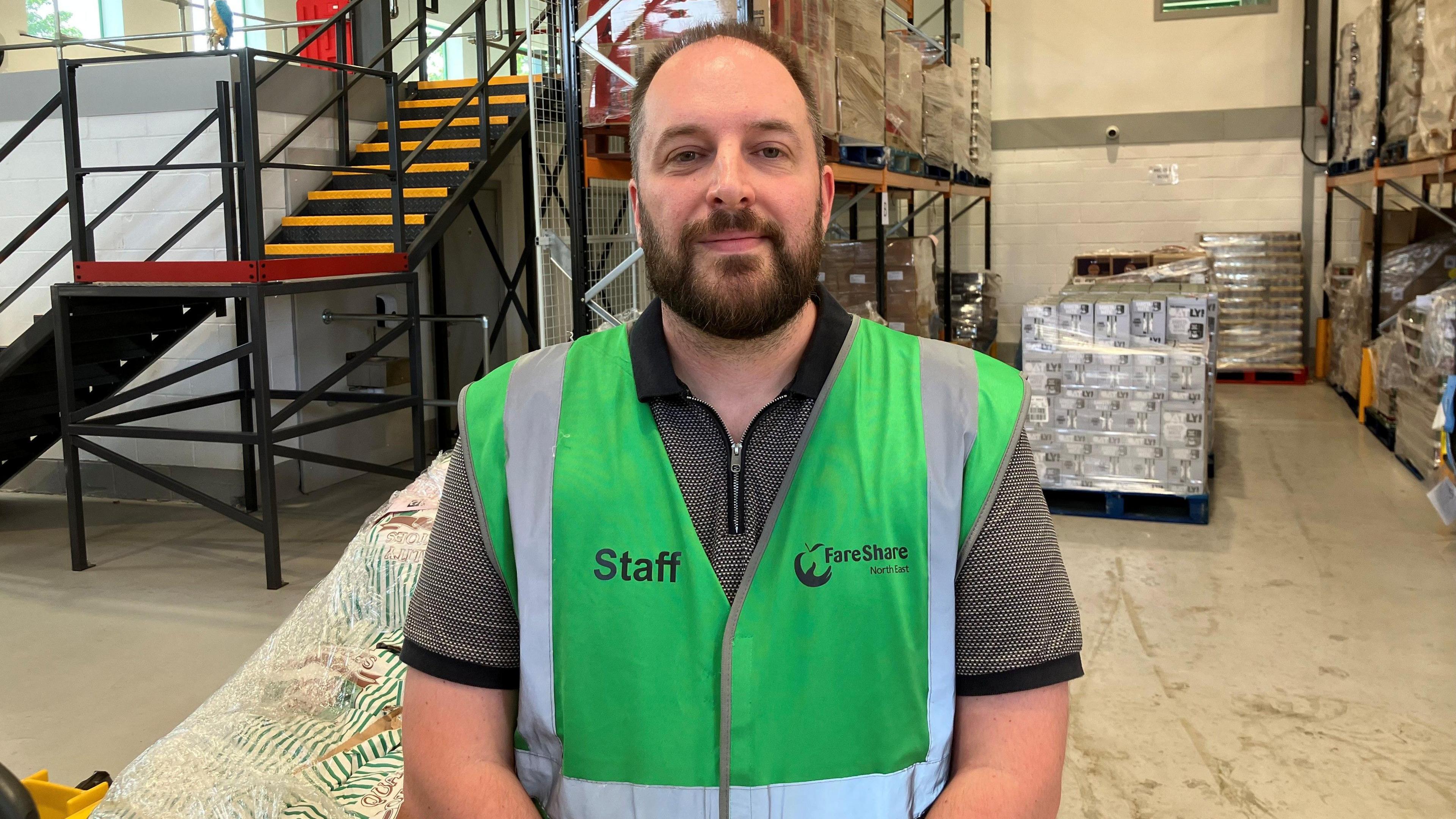 Jake Hanmore, CEO of FareShare North East, standing inside the Middlesbrough hub. He is wearing a grey t-shirt and a green vest with the charity's logo. He has short dark hair and a beard. 