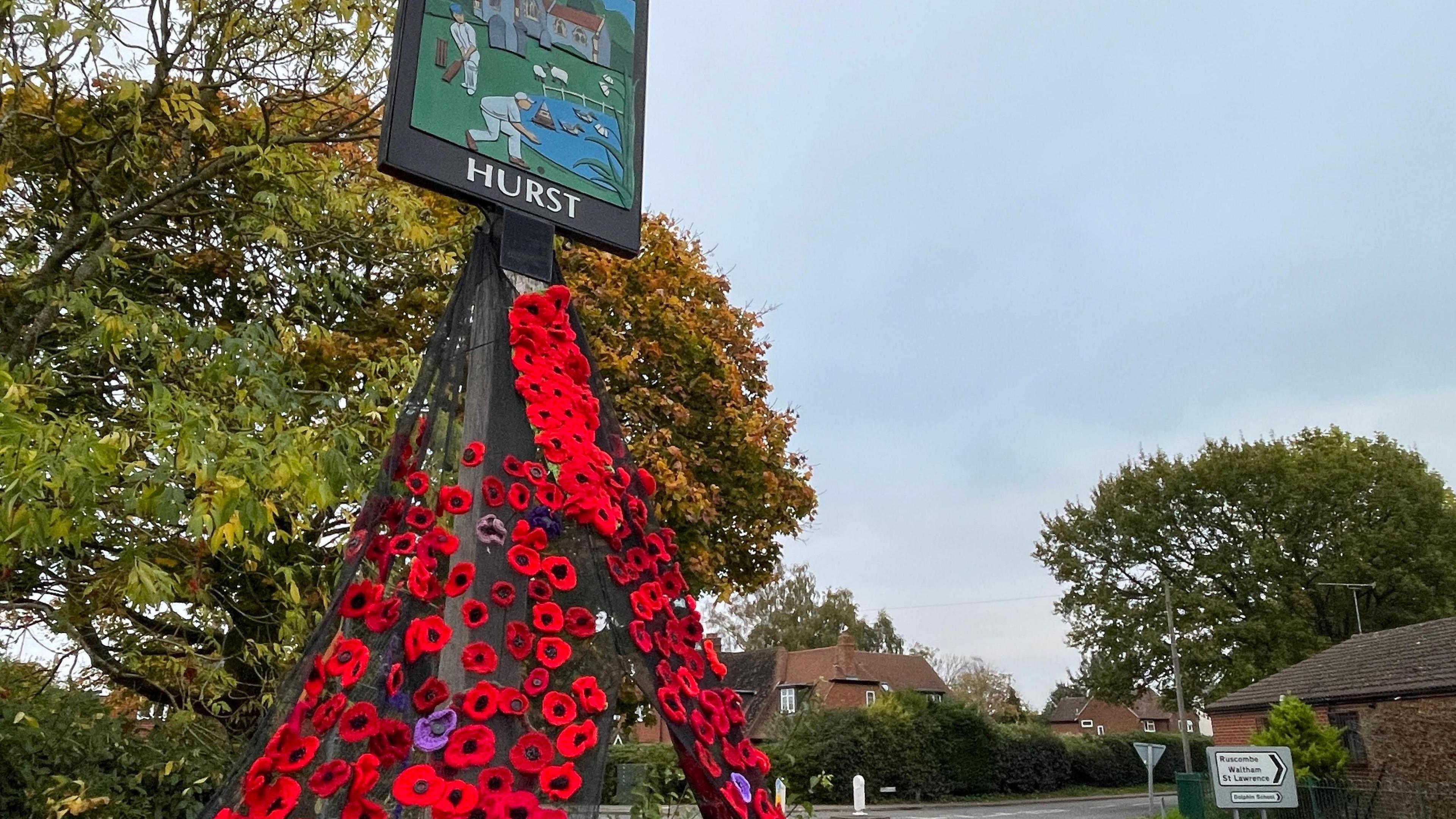 A number of red knitted poppies, with a few that are purple, cascade downwards from a village sign. There are trees and a road in the background of the shot. The sign says 'Hurst'.