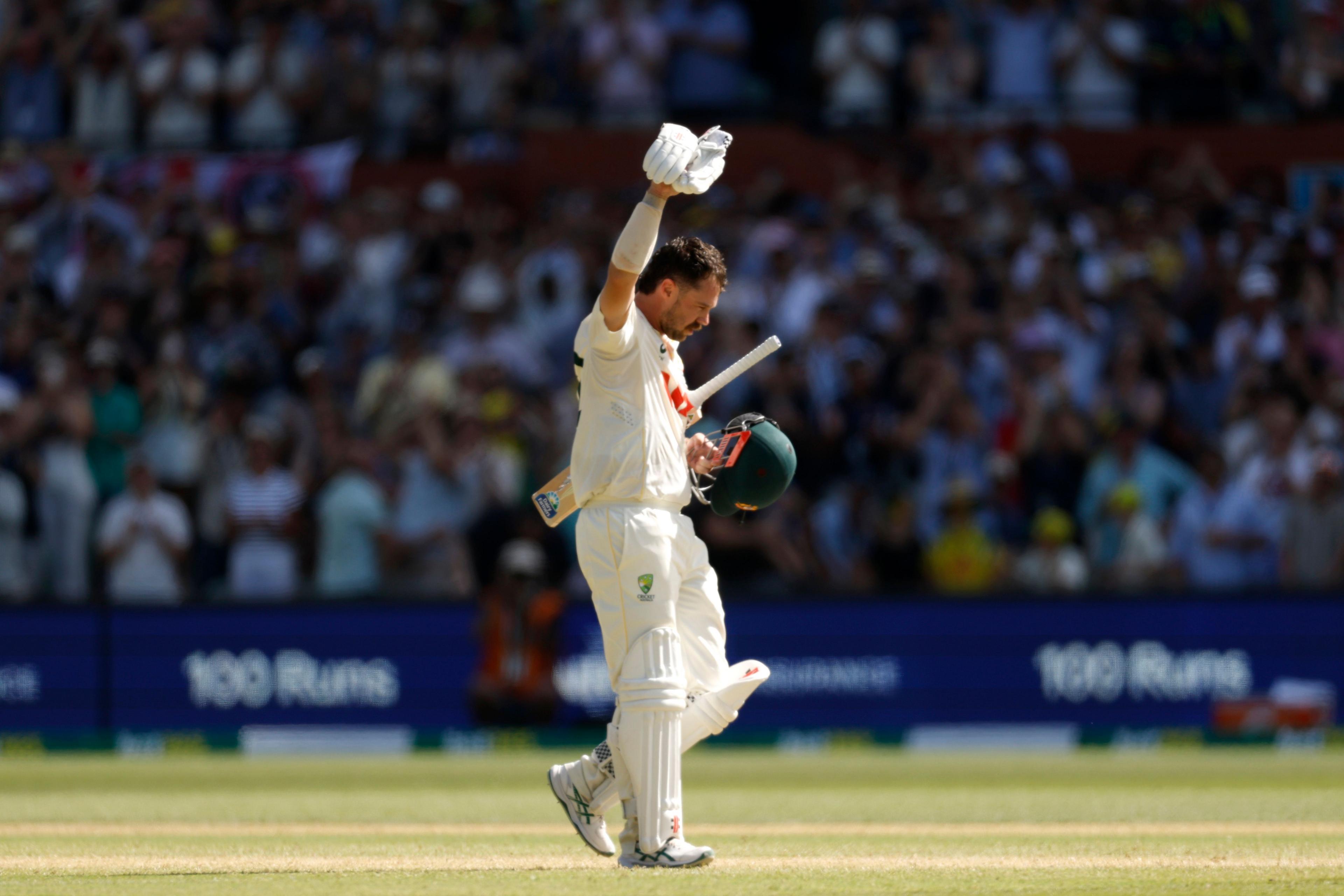 A cricket player in white kit walks on the field raising their bat and helmet in celebration, acknowledging the cheering crowd in the background. The stadium is full, and a scoreboard display behind them shows “100 Runs,” indicating a milestone achievement.