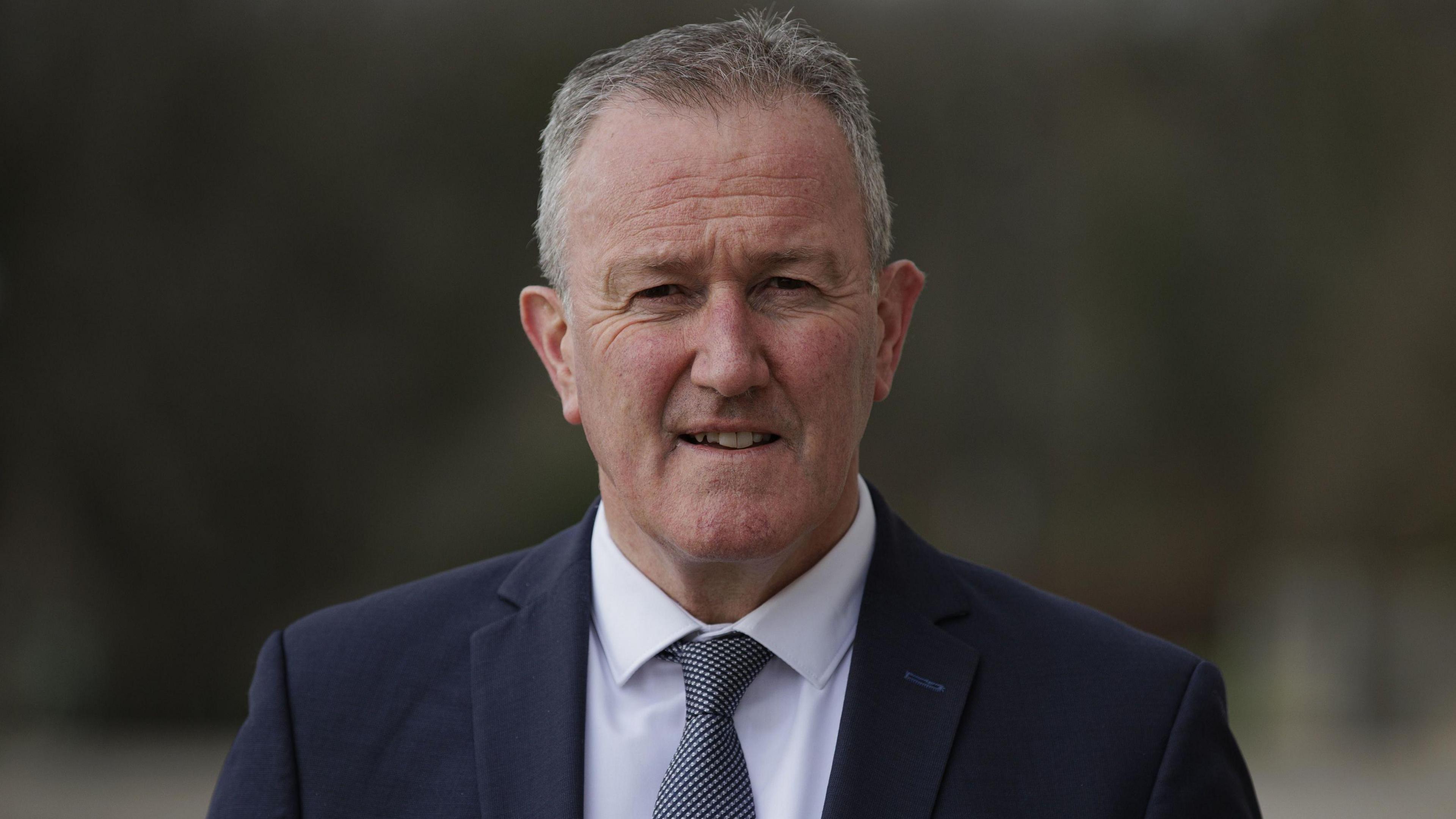 A man with grey hair smiles in the direction of the camera. He is wearing a dark navy suit with a white shirt and a grey and white tie.