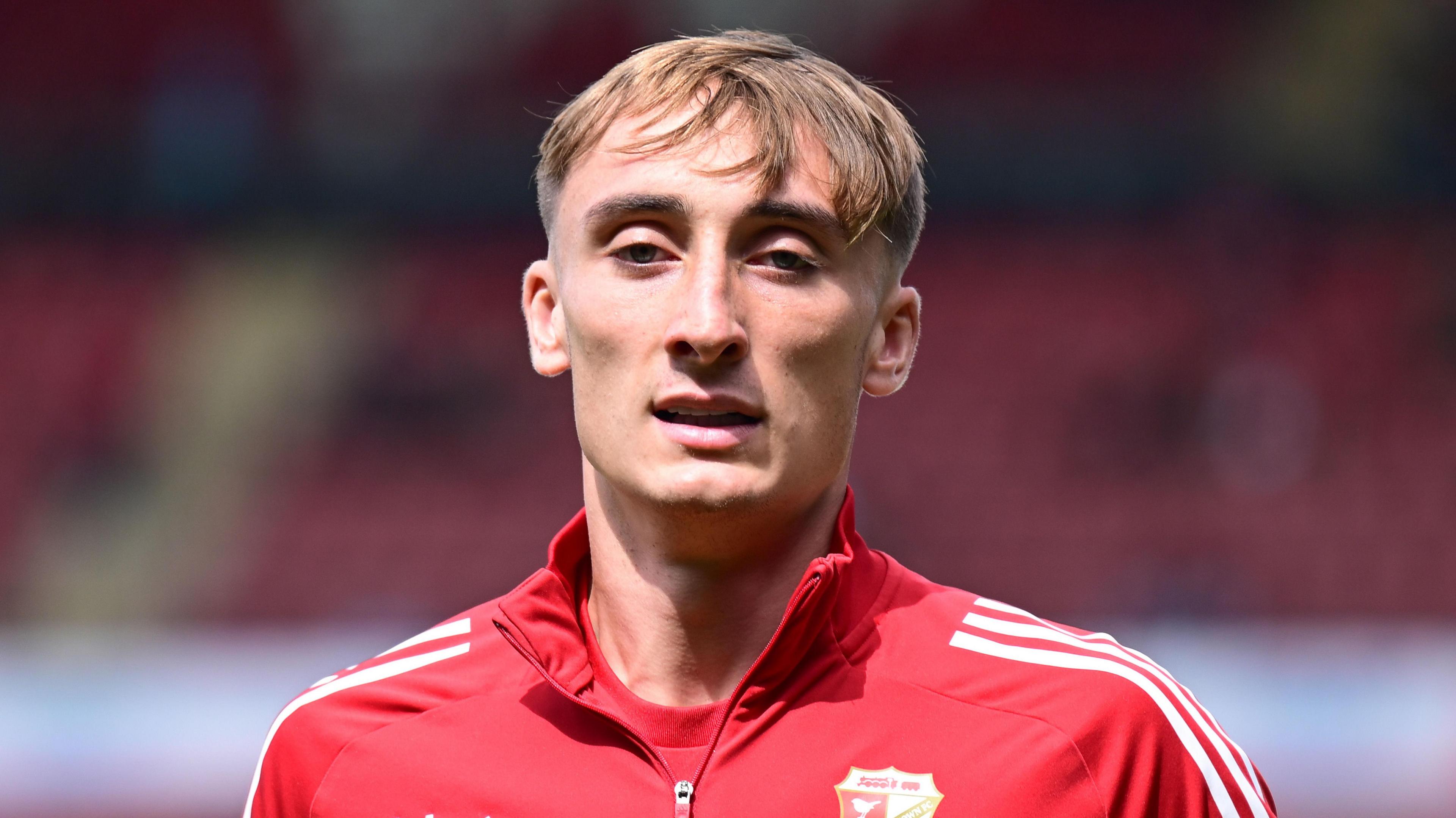 Jake Tabor wearing a red Swindon Town jacket and standing on the pitch during a pre-game warm-up in August 2025.
