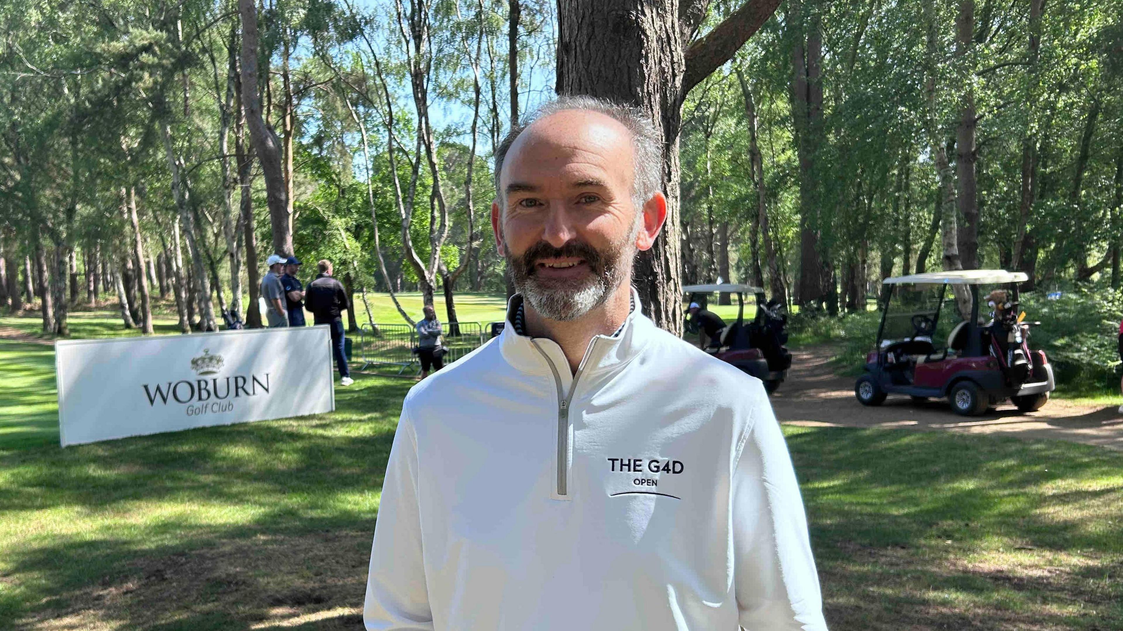 Mr Barker is standing on the golf course wearing a white 3/4 zip jumper with green branding on the right breast pocket. It reads The G4D open. He has grey and dark hair and facial hair. He is smiling at the camera. behind him are some golf carts.