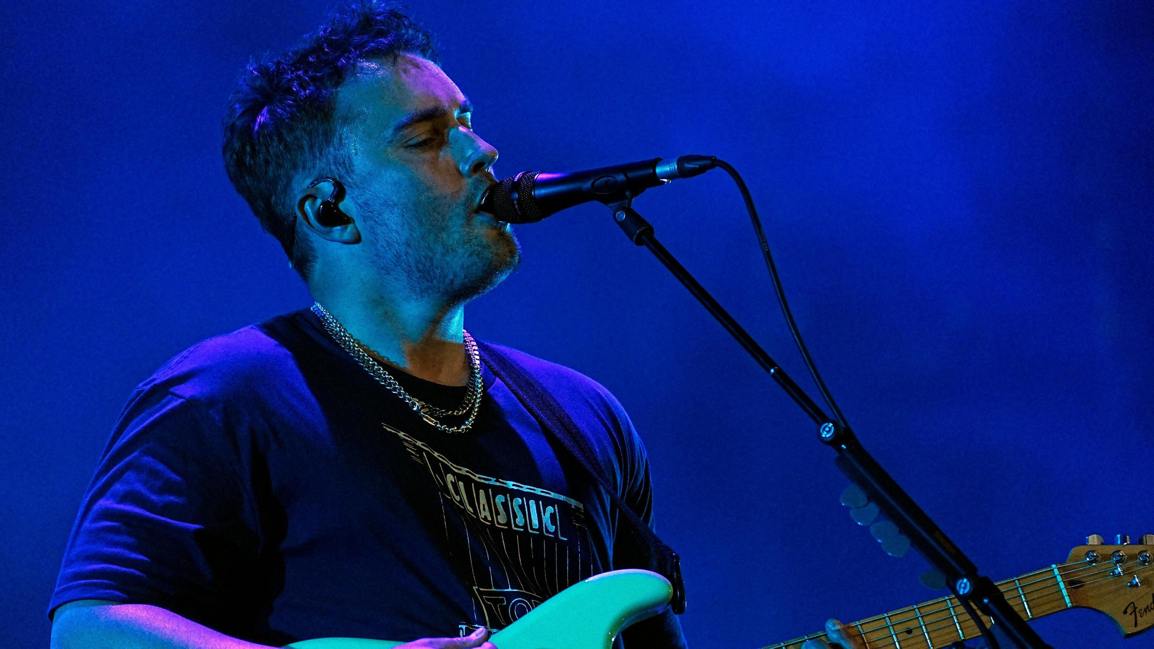 Sam Fender wearing a T-shirt, holding a white electric guitar performing in front of a microphone stand. 