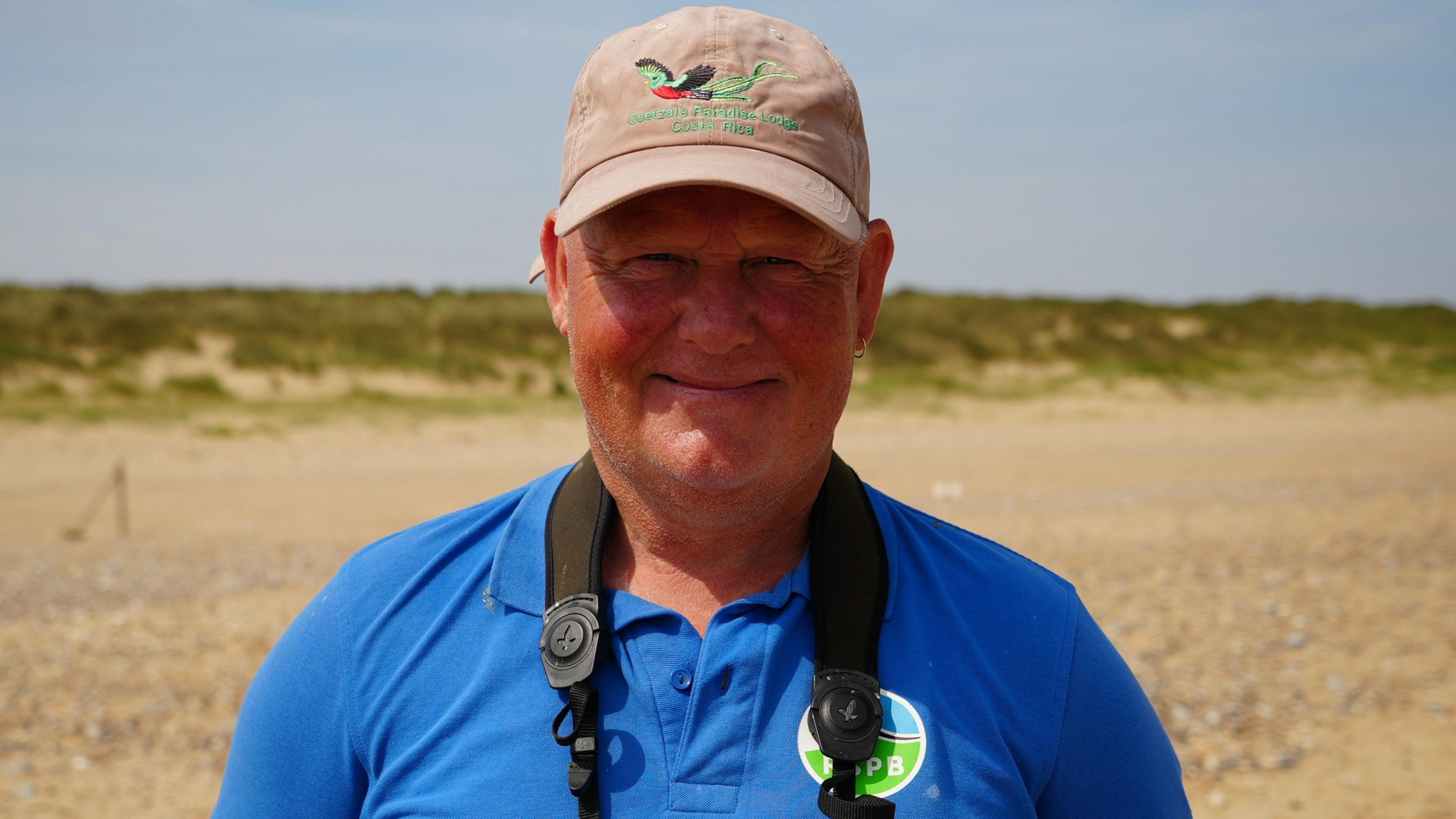 A man smiling at the camera in a head and shoulders image. He is wearing a blue polo shirt and a beige baseball cap.