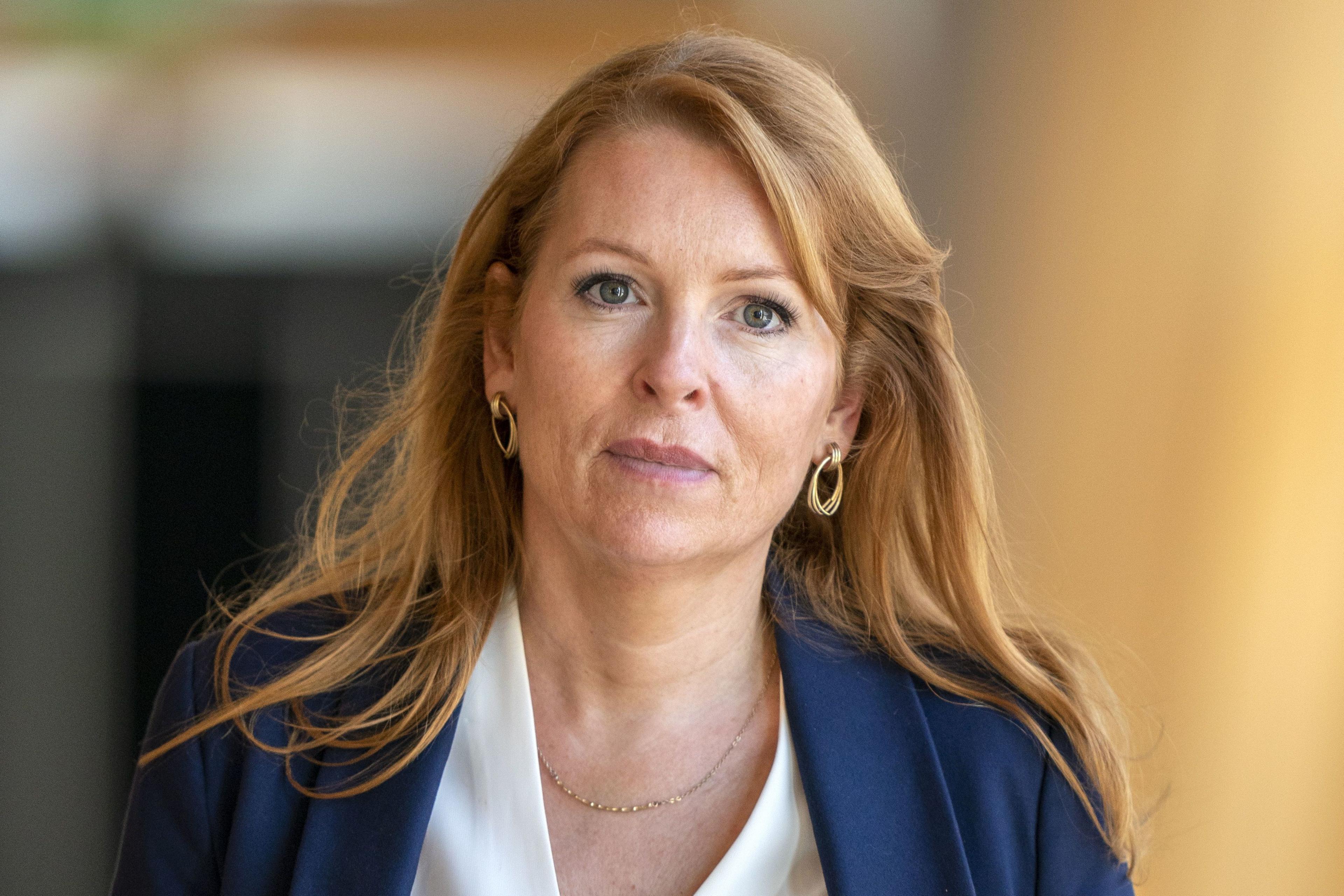 Ash Regan MSP in the Scottish parliament. She is wearing a blue jacket, white blouse and gold ear rings. She has long reddish hair and is smiling. the background is out of focus.