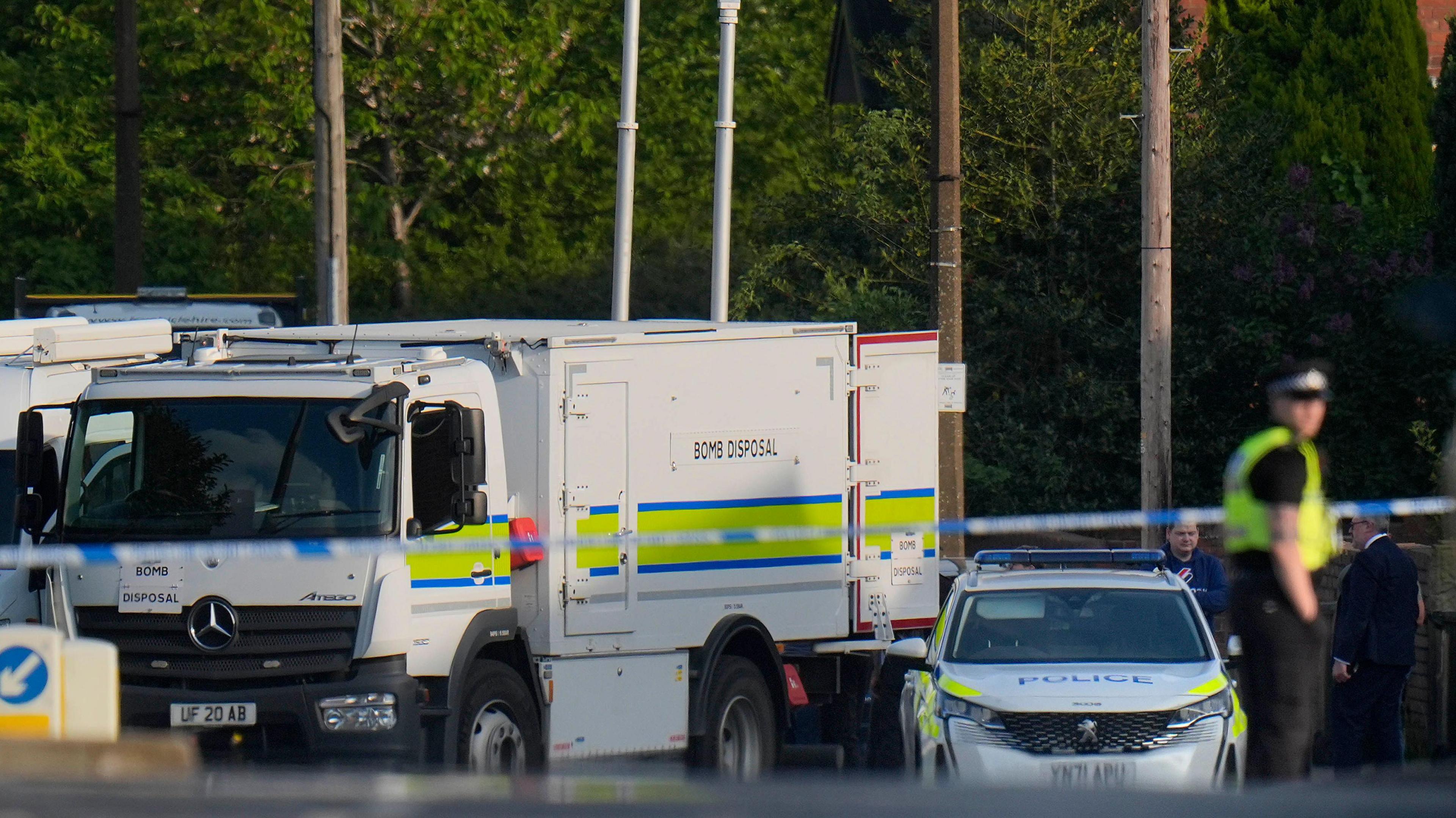 A scene involving emergency response vehicles and a secured area. A white bomb disposal truck with blue and yellow stripes is prominently visible, clearly marked with the words "BOMB DISPOSAL" on its side. Next to it is a white police car with matching blue and yellow markings. The area is cordoned off with police tape.
In the background, several individuals can be seen, including at least one person wearing a high-visibility vest.