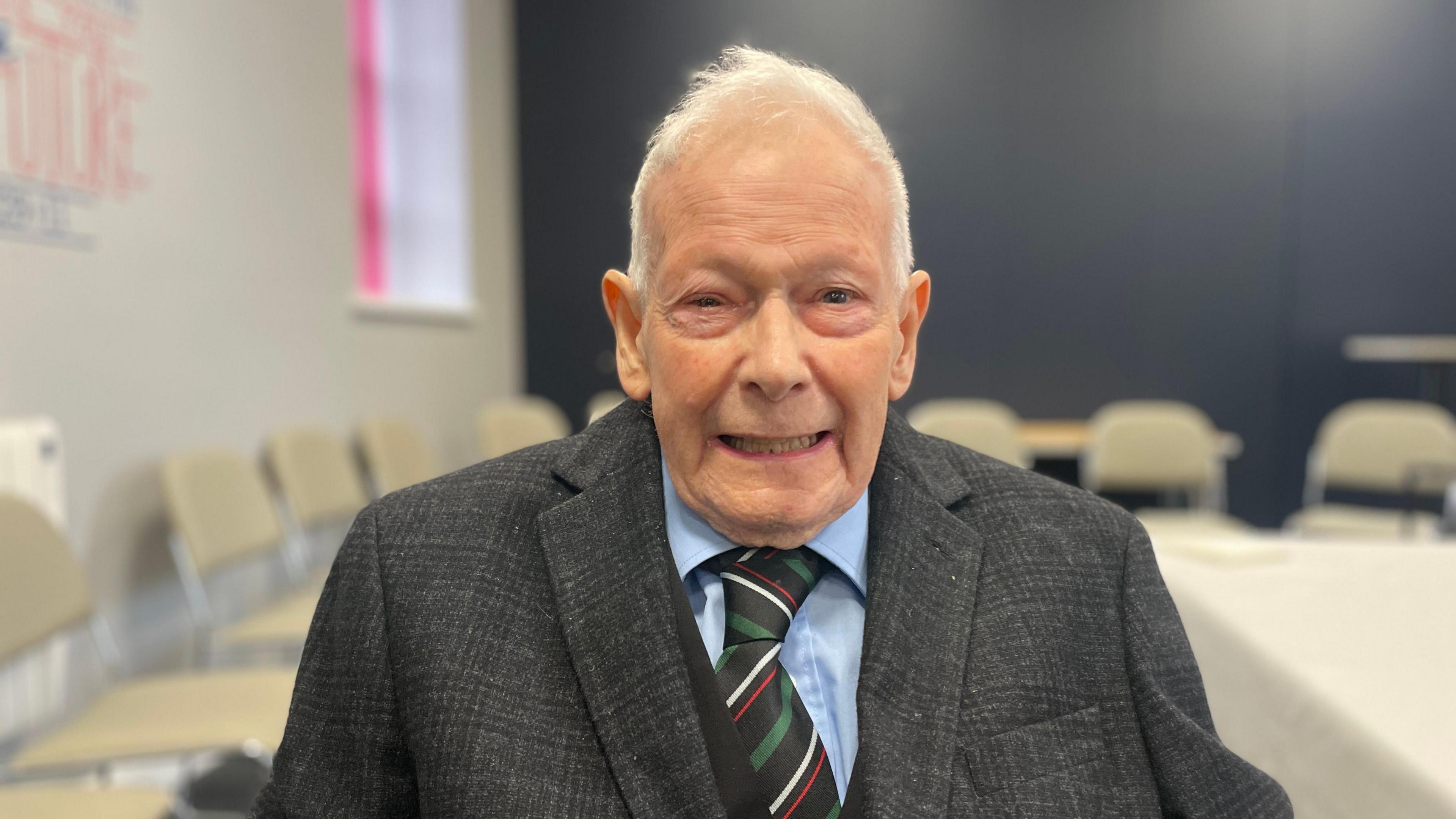 Reverend Robert Hanna looking directly at the camera and smiling. He has white hair wearing a dark grey blazer, light blue shirt, and green/red/white/black striped tie. 