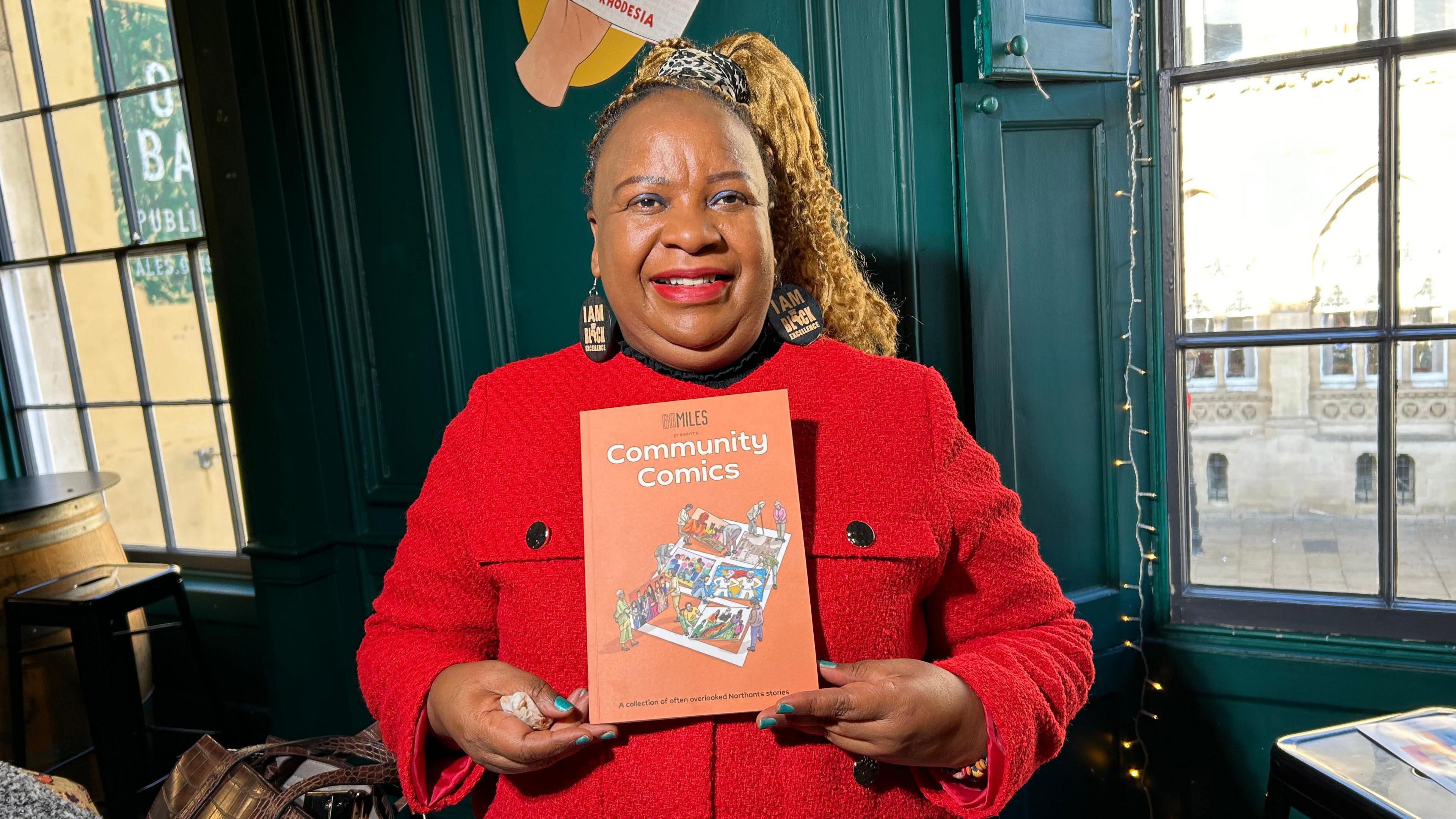 A woman standing in a room with large windows and wooden panelled walls. She is wearing a bright red winter coat and holding up an A4 sized book which is titled 'Community Comics'