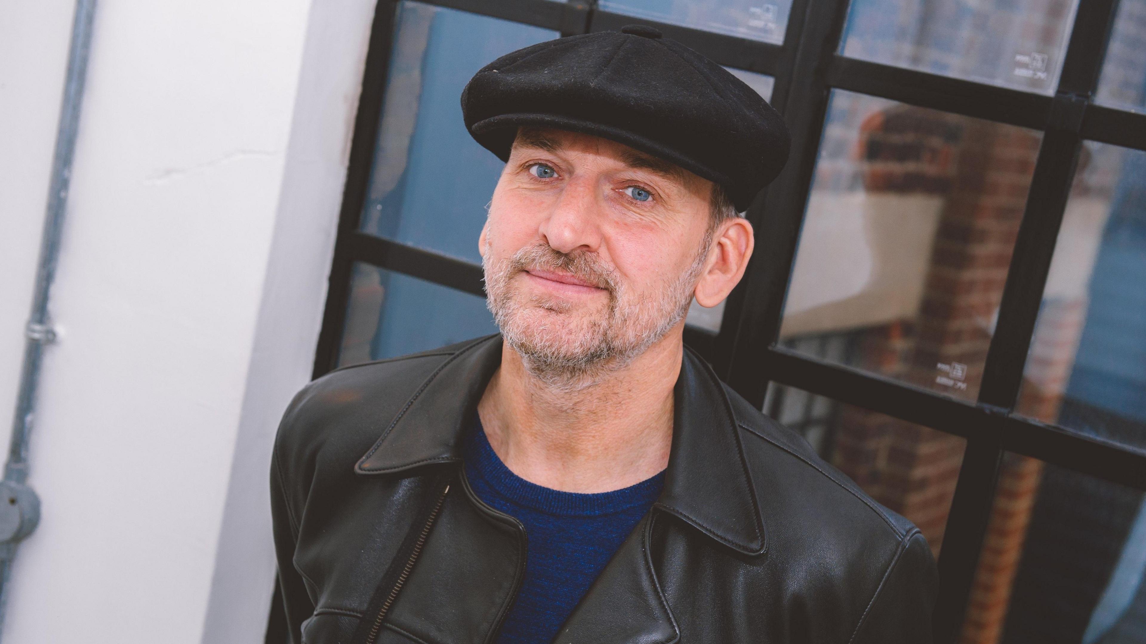 A man in a peaked cap and leather jacket looks at the camera.