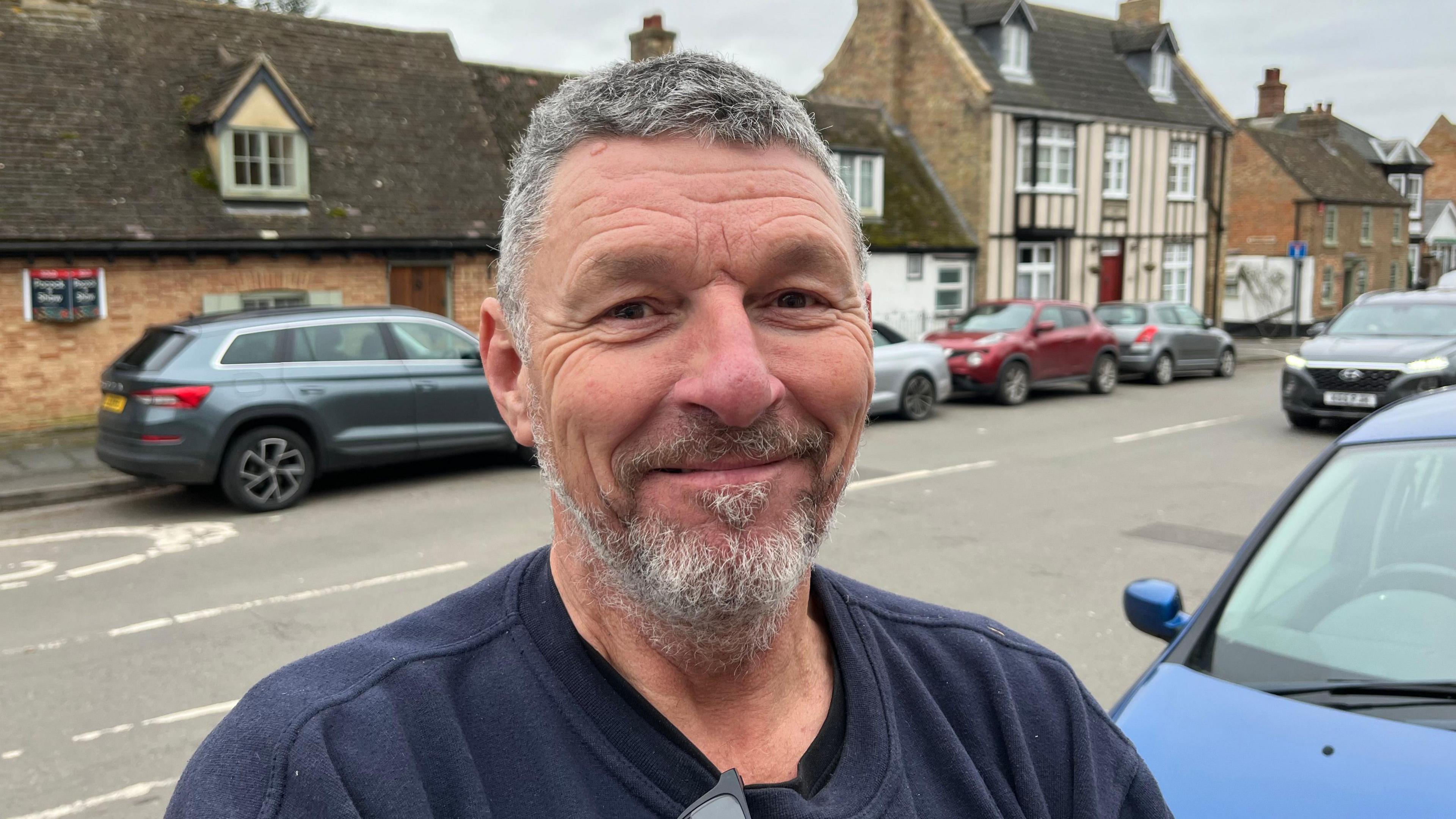 A smiling Kevin Woolridge looks directly at the camera as he is photographed outside on a street pavement. He is wearing a navy jumper with sunglasses hanging from his neckline. There is a row of houses behind him and a number of cars parked on the street. 