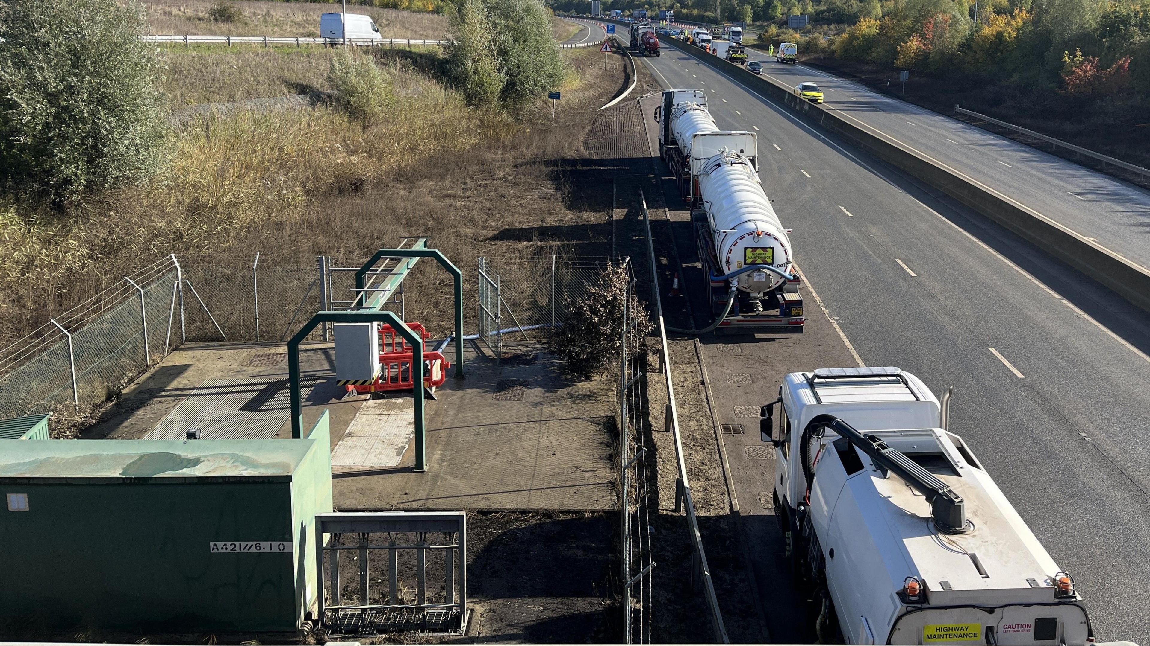 Three tankers can be seen parked at the side of the A421 next to a pumping station. They have long hoses that are stretched to the pumping station. Cars can be seen in the distance on the carriageway.